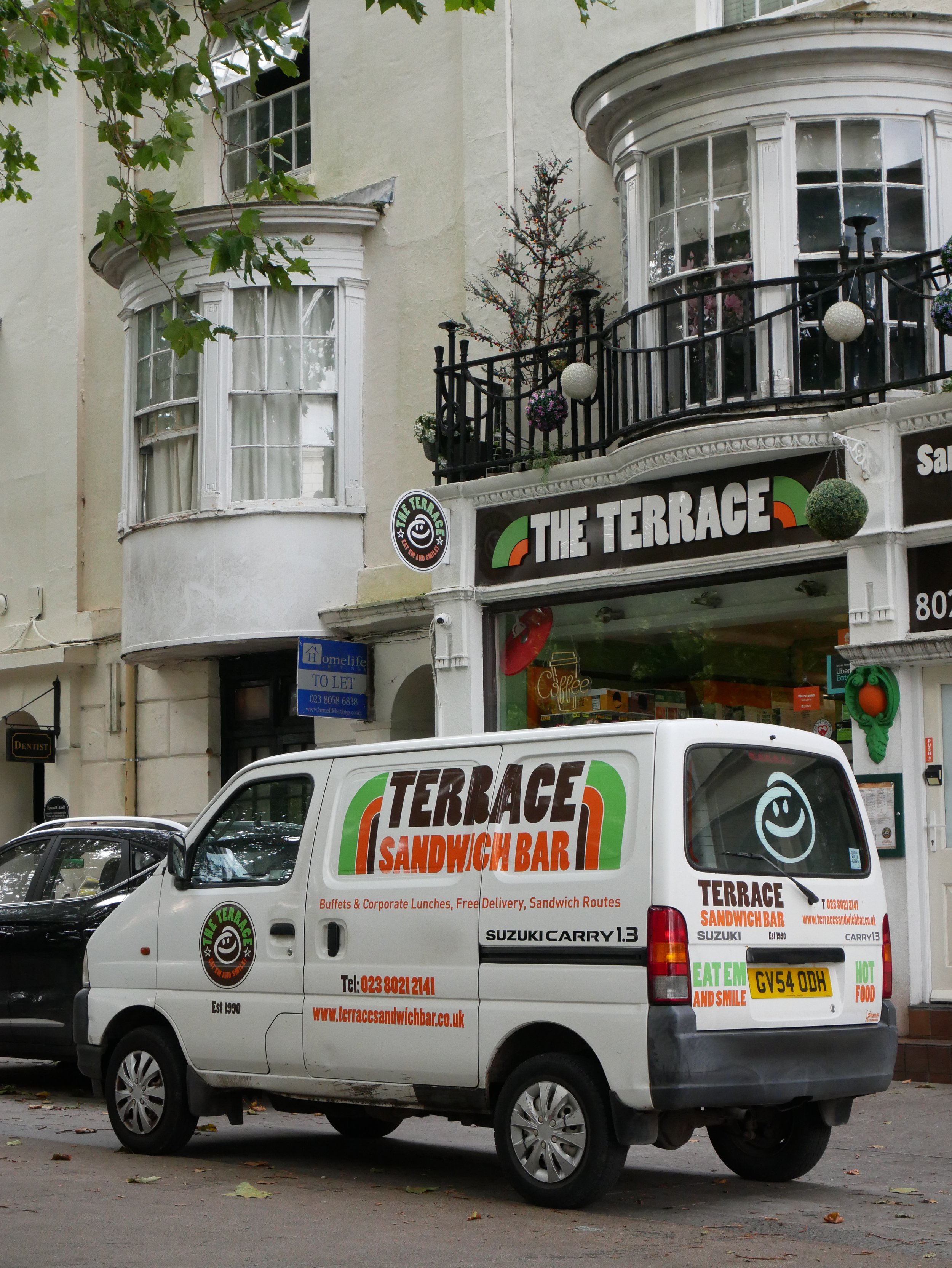 A white van with signage for Terrace Sandwich Bar, parked on a street. In the background, there is a building with bay windows and a balcony decorated with potted plants. The van displays the restaurant’s logo, contact information, and menu highlight