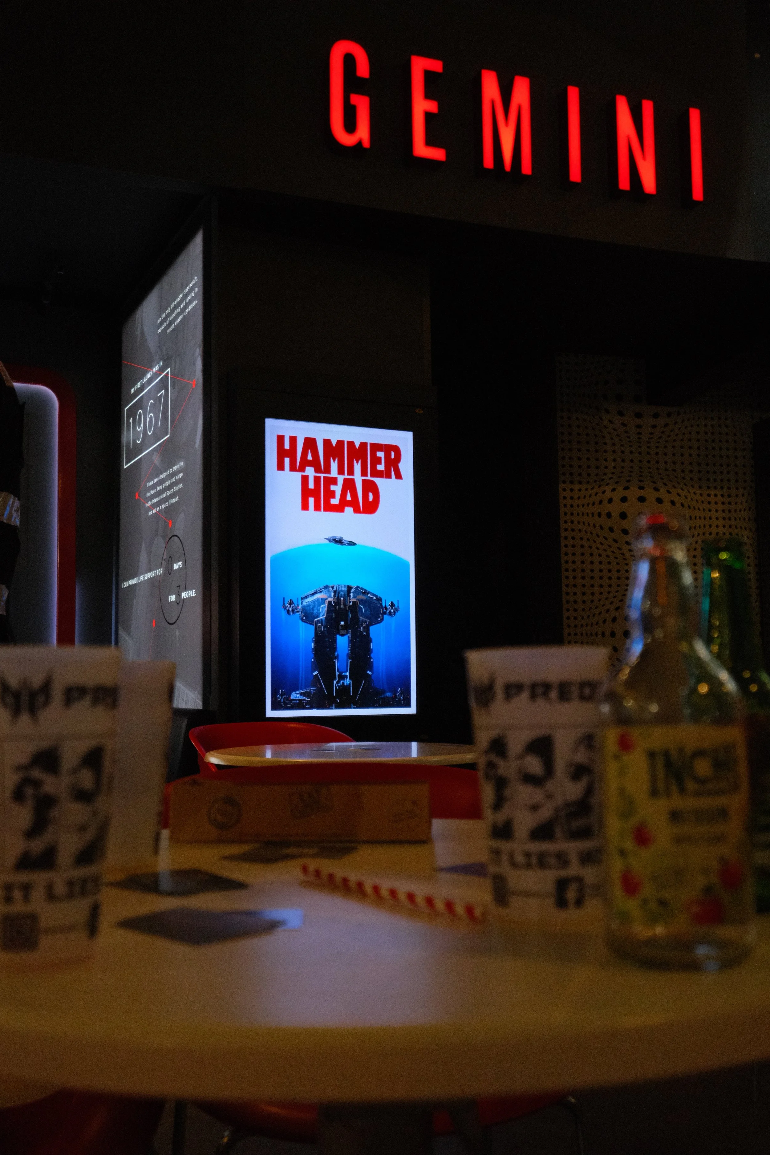 The image shows a dark interior space with a large red illuminated sign that says 'GEMINI'. There is a poster advertising 'HAMMER HEAD' with a picture of a robotic vehicle. The foreground contains out-of-focus objects, possibly food packages and bott