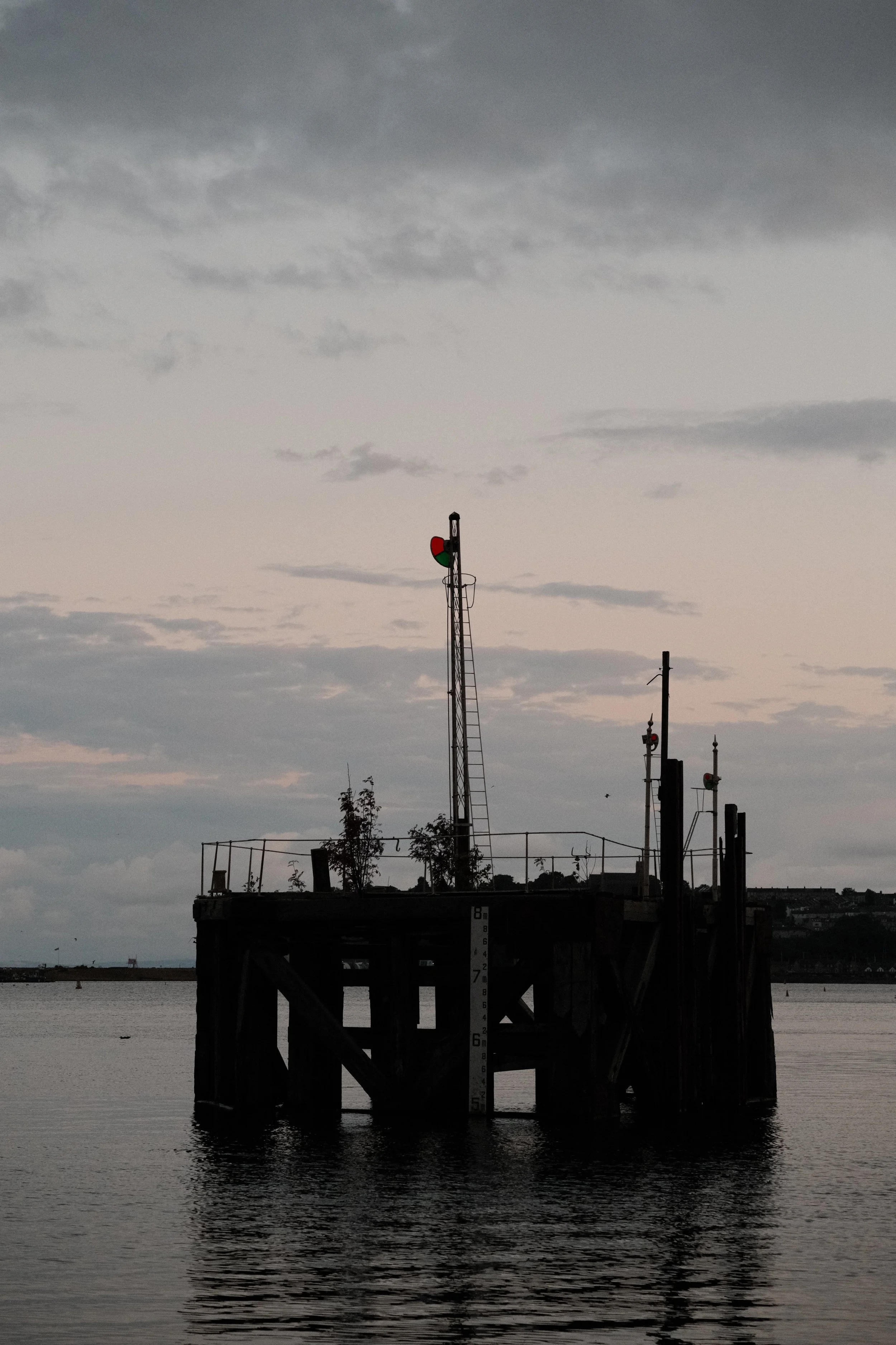 A silhouette of a pier with navigation lights and a weather monitor, with water and a cloudy sky in the background, near sunset or dusk.