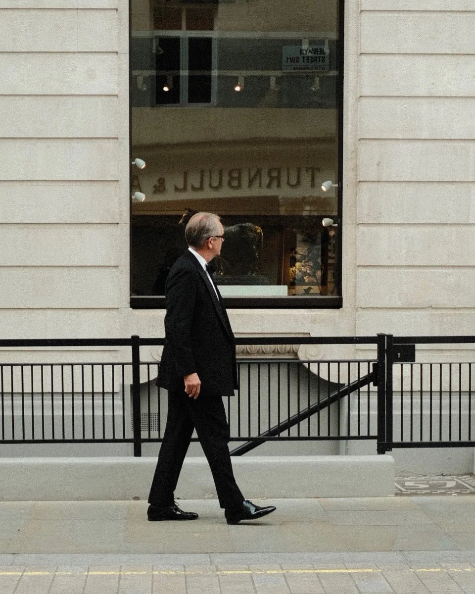 A man in a black suit and glasses walking on a city sidewalk with a building's window and a railing behind him.
