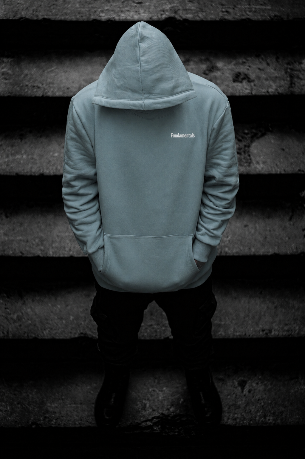 Person standing with head down, wearing a light-colored hoodie with the word "Fundamentals" on it, and black pants, against a dark textured background.