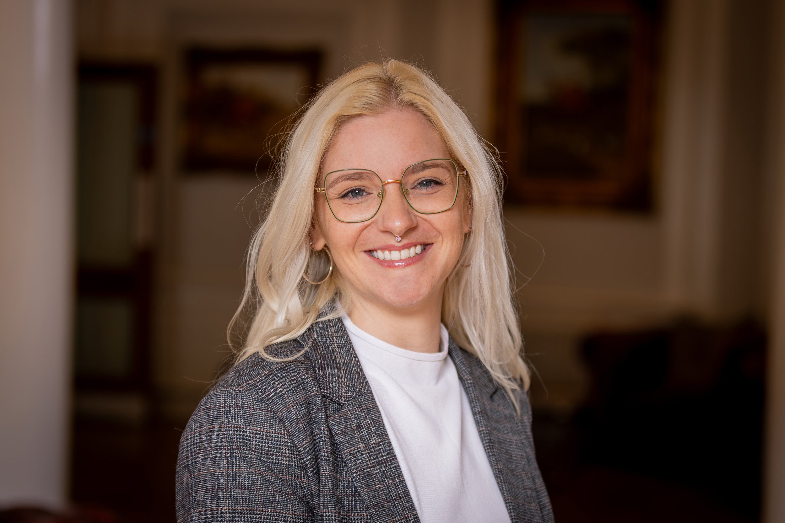 A woman with long blonde hair and glasses smiling, wearing a plaid blazer and white shirt, standing indoors with a blurred bookshelf or shelving in the background.