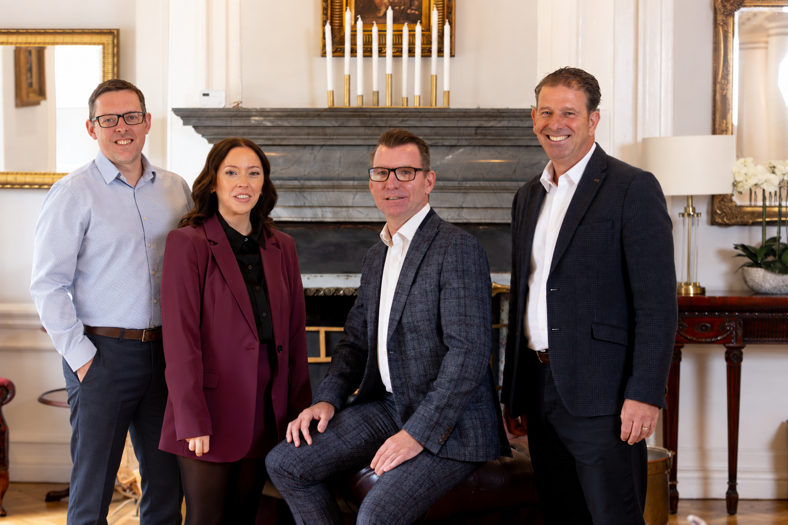 Four professionally dressed individuals, three men and one woman, standing and sitting in a formal room with a fireplace, mirror, and gold accents, smiling at the camera.