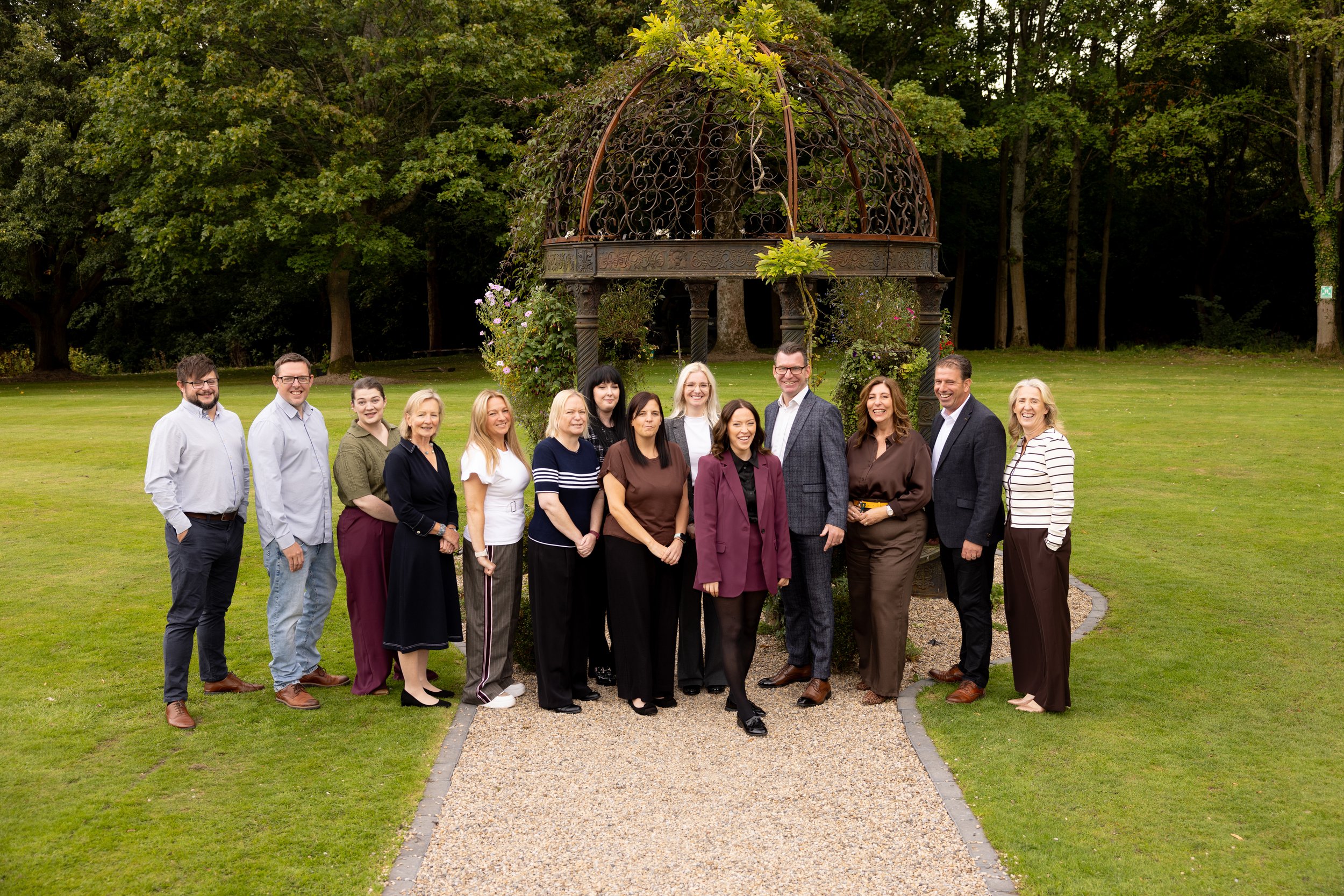 A group of sixteen people standing outdoors on a grassy area, posing for a photo in front of a decorative metal gazebo with surrounded by trees.