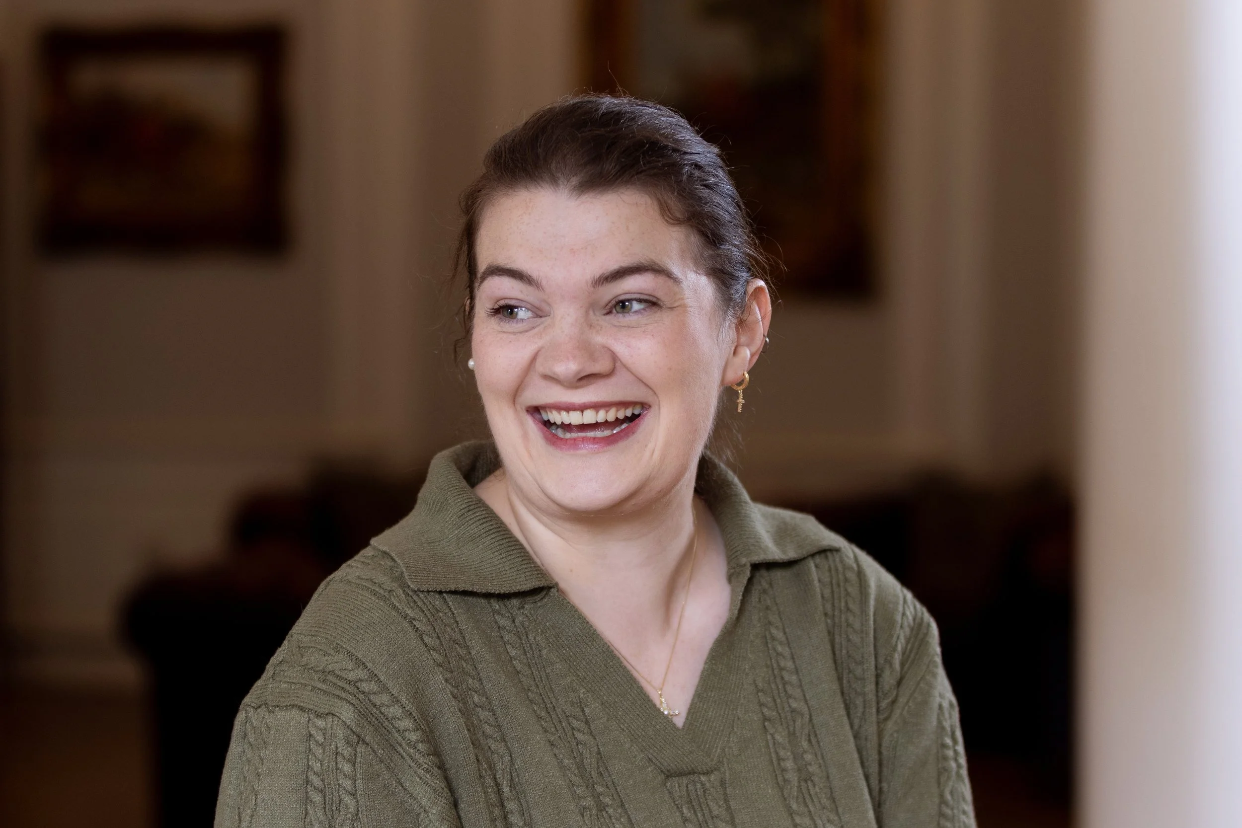 A young woman with dark hair pulled back, smiling and looking to her left, wearing a green knitted sweater, in an indoor setting with blurred artwork in the background.