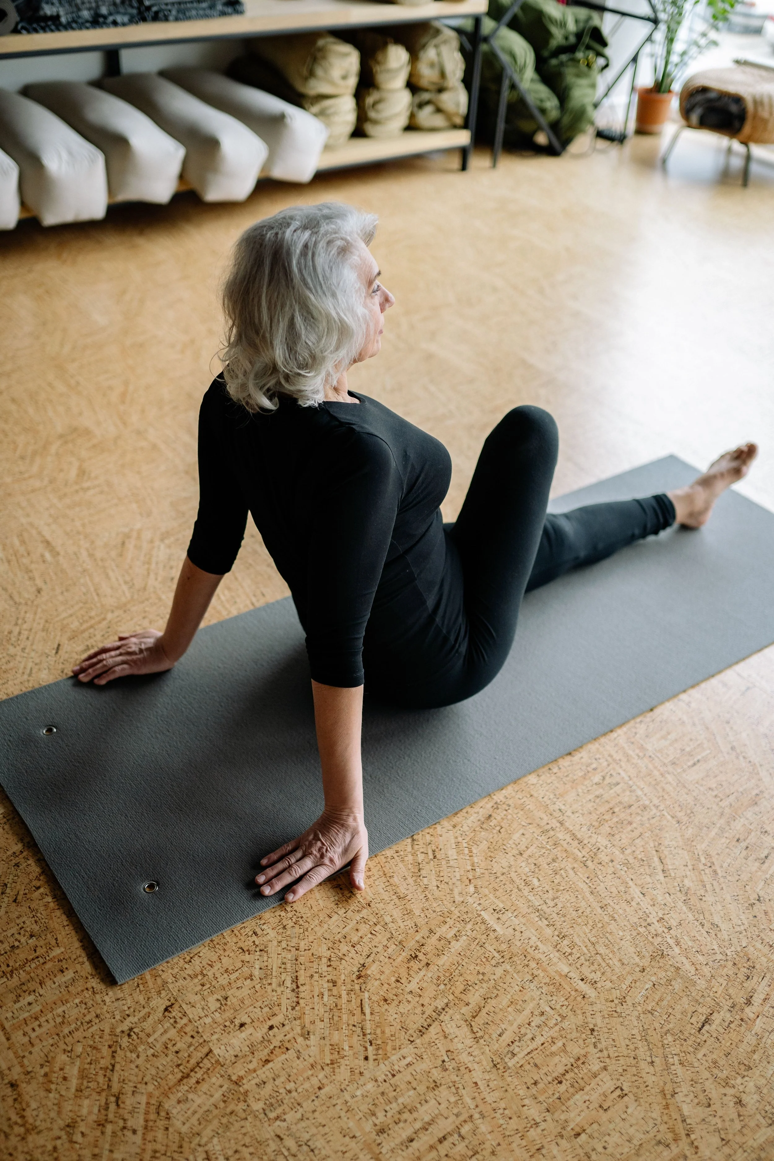 Older woman practicing yoga on a mat in a cozy indoor space with wooden floor, shelves with bags, and potted plants in the background.