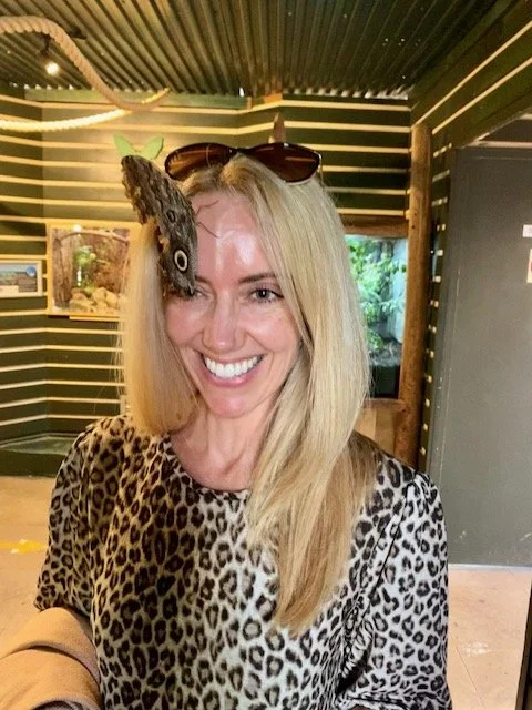 A woman with blonde hair smiling, wearing animal print clothing and sunglasses on her head, has a butterfly and a small green leaf on her forehead.