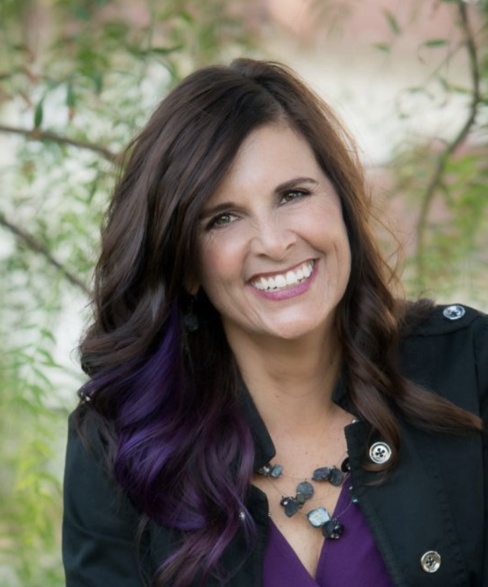 A woman with long, wavy dark hair with purple tips, smiling outdoors with green foliage in the background.