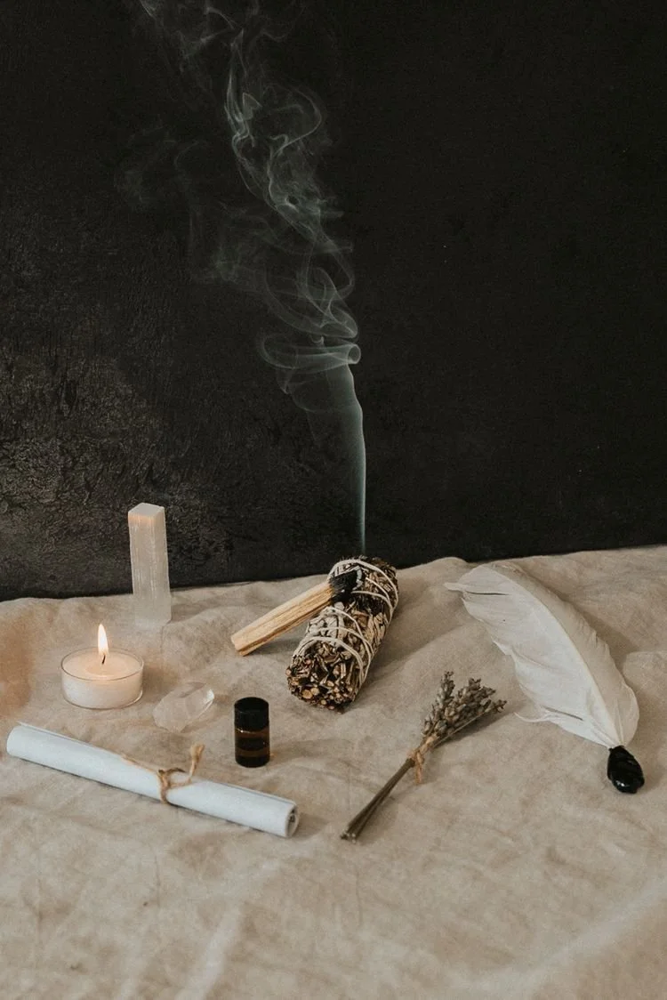A spiritual or ritual setup with a lit candle, incense sticks, a bundle of sage, a small bottle of oil, a quartz crystal, a white feather, and a bundle of herbs on a cloth surface against a dark background with smoke rising.