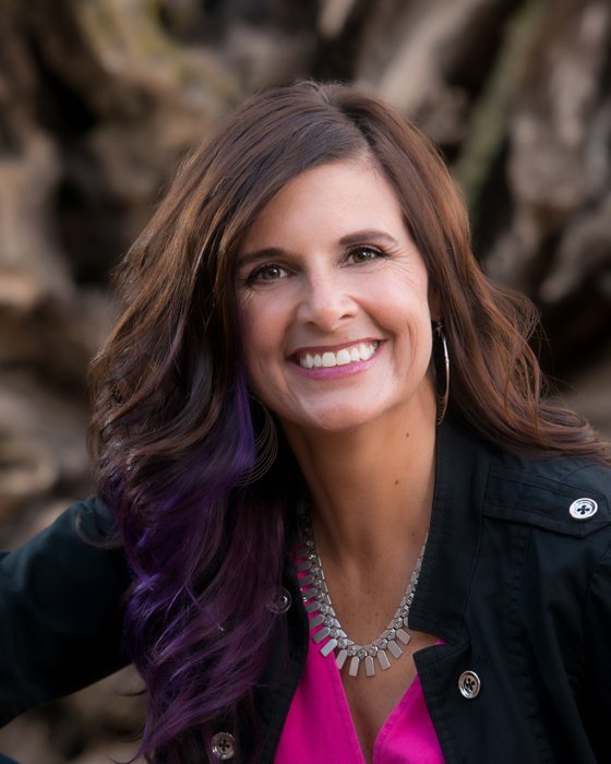 A woman smiling outdoors with long brown hair that has purple streaks, wearing a black jacket, pink top, and silver necklace.
