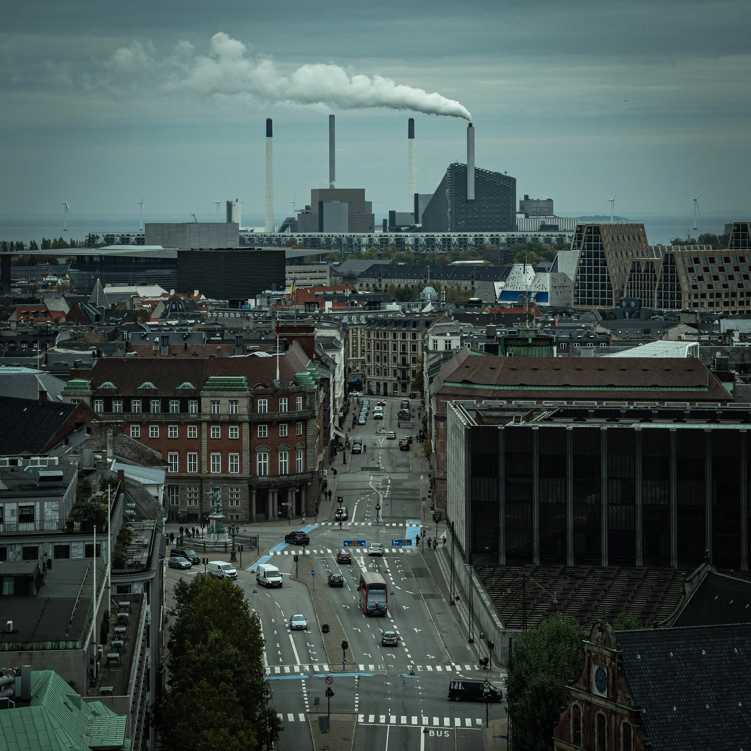 View of a city street intersection with cars, buses, traffic lights, and buildings, leading to a large industrial complex with smoke stacks emitting smoke from CopenHill (also known as Amager Bakke), wind turbines in the background, and cloudy sky.