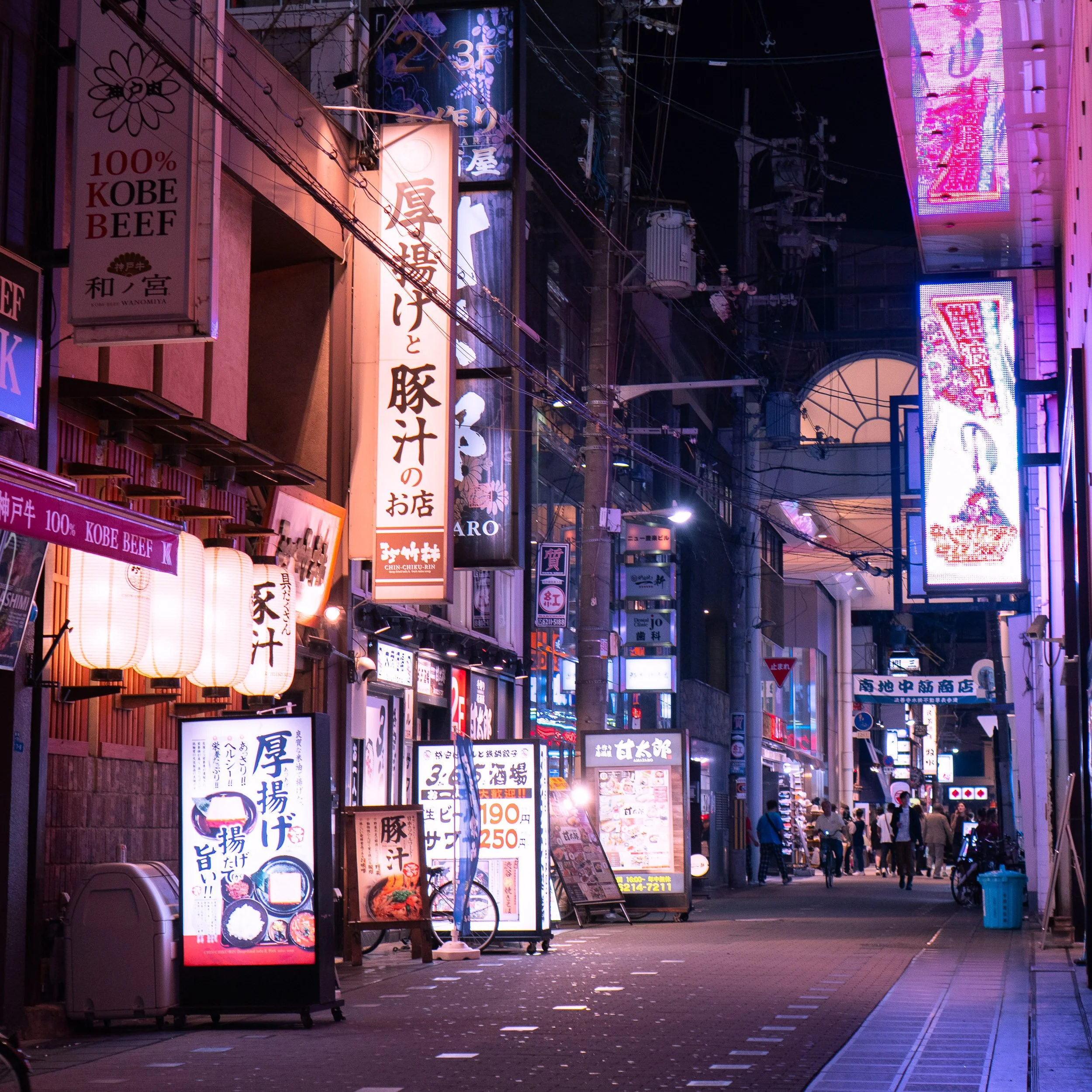 Nighttime street scene with illuminated signs in Japanese, paper lanterns, and people walking on the sidewalk.