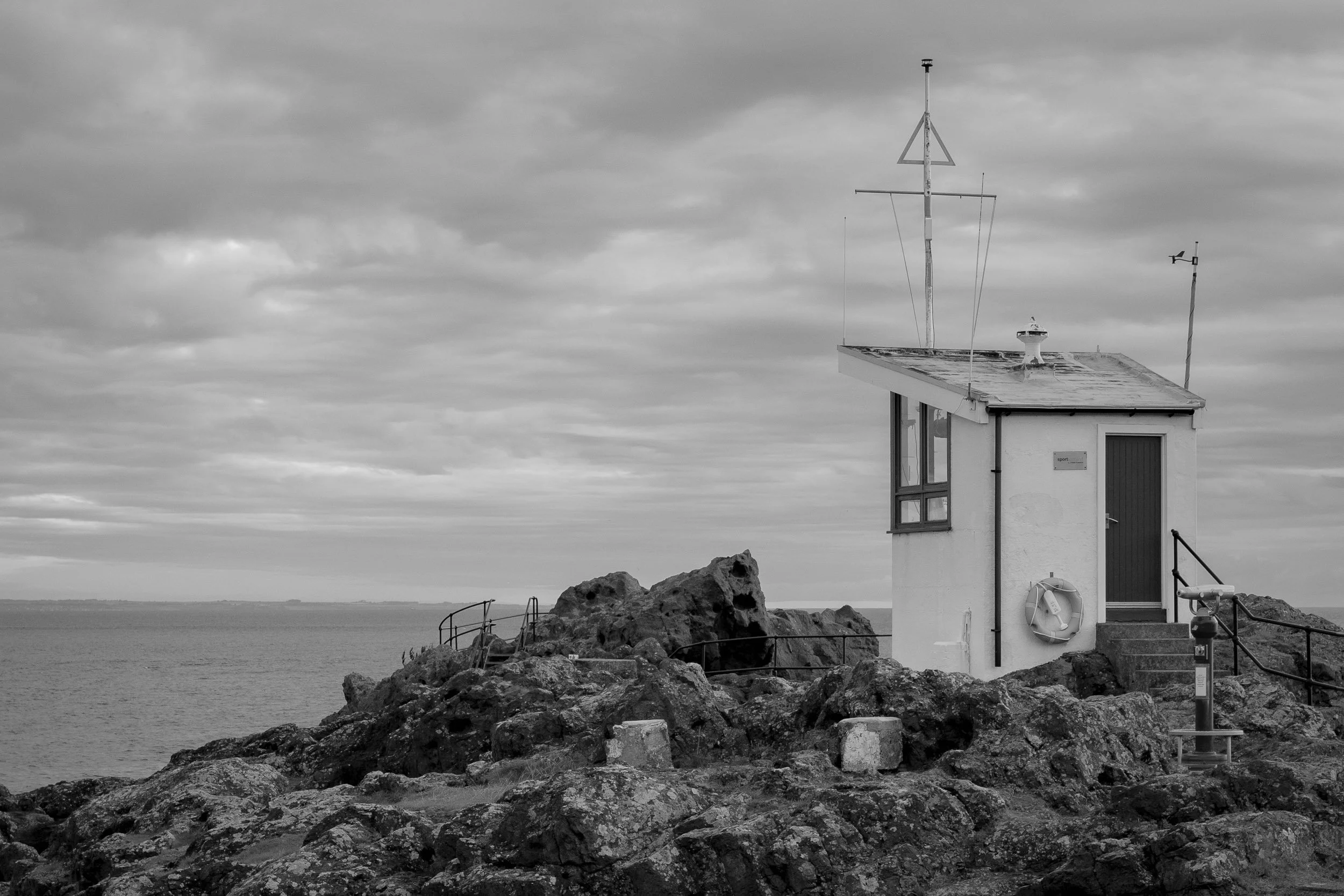 A black and white photo of a small lighthouse or weather station on a rocky coast, with antennas on the roof, among rocks, with a dock or pier and water in the background under a cloudy sky.