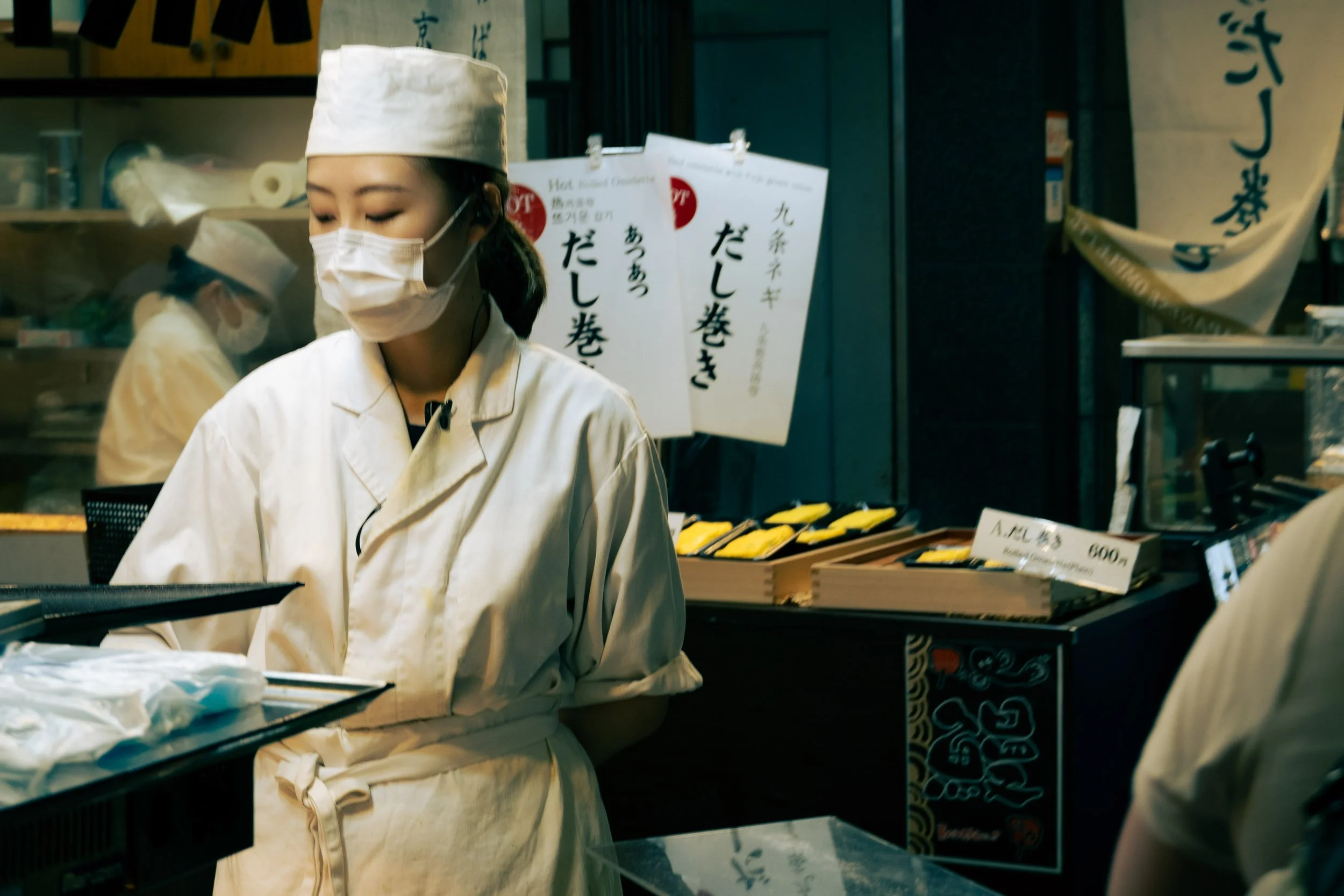 A woman wearing a white chef's coat, face mask, and head covering, working at a food stall or preparation area. In the background, there are signs and a display with Japanese food items, with prices and descriptions.