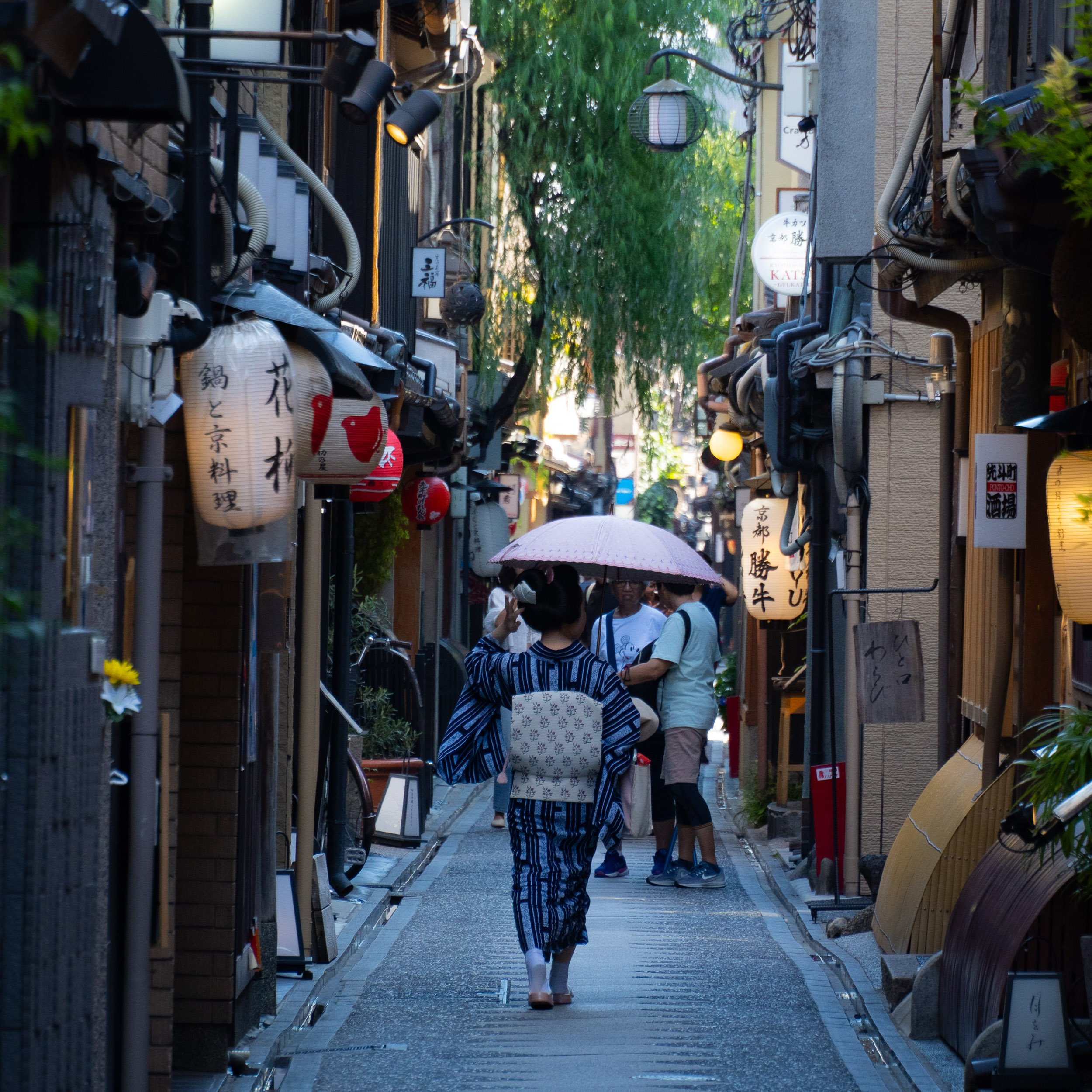 A woman in traditional Japanese clothing walking down a narrow street in Kyoto with hanging lanterns, signs with Japanese characters, and lush greenery in the background.