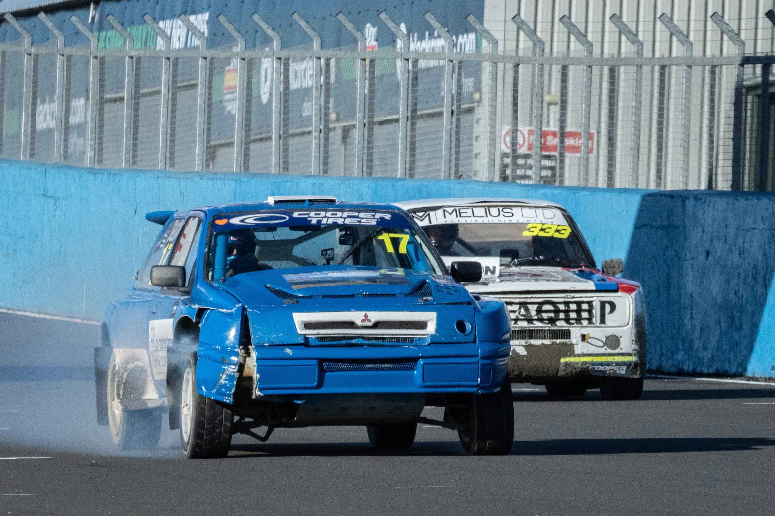 Race cars are racing in a Rallycross event at Knockhill, Scotland, with visible tire smoke resulting from damage to the blue car.