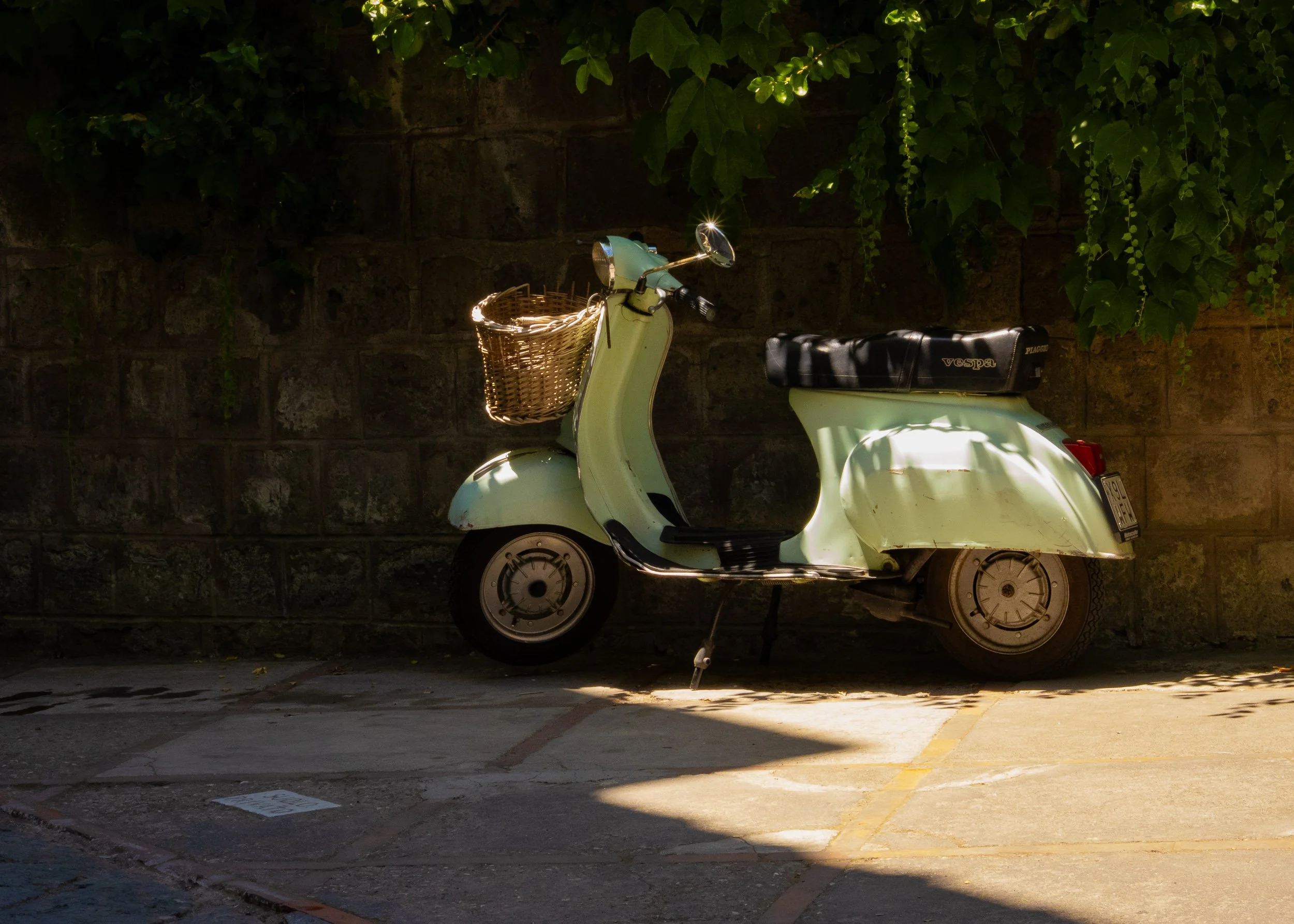 A vintage pale green Vespa scooter with a wicker basket attached to the front, parked on a street in Sorrento with a stone wall and green ivy in the background, partially illuminated by sunlight.