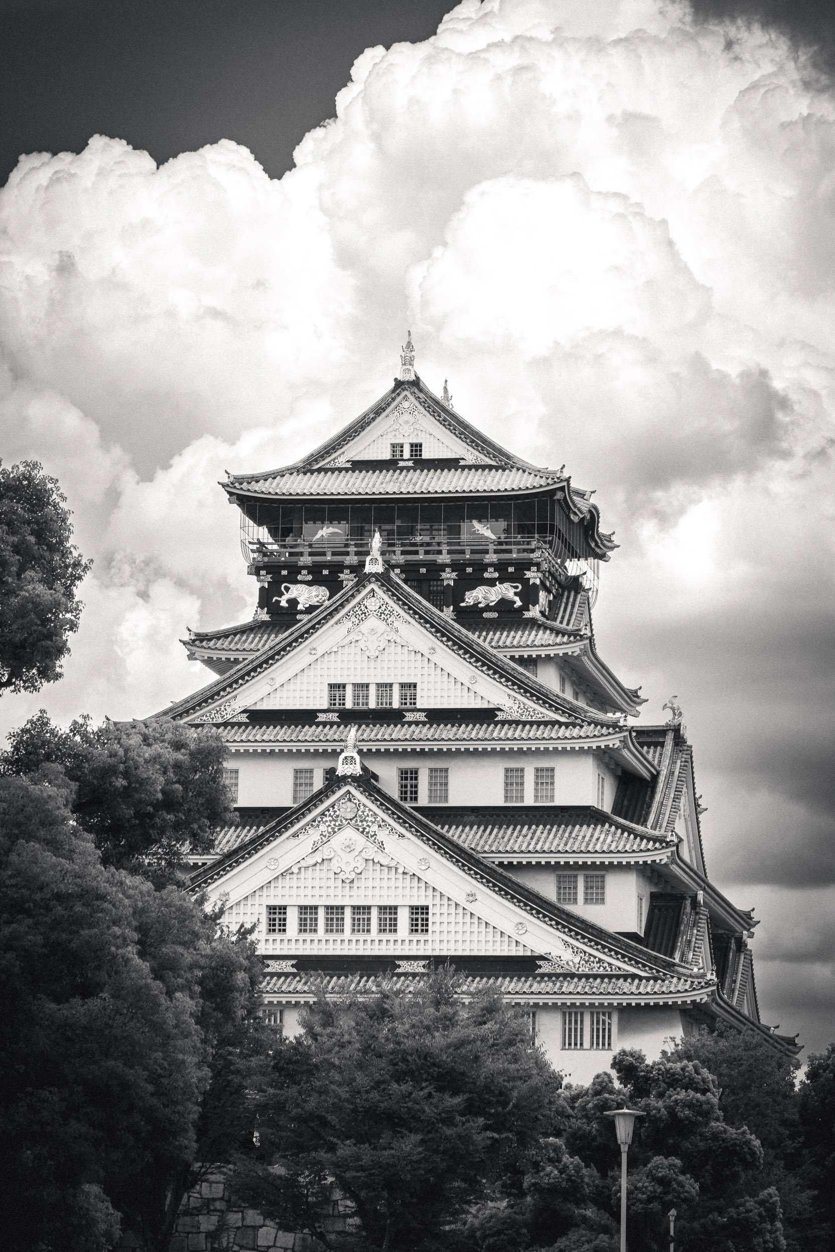 Osaka Castle with multiple layered roofs and intricate architectural details, surrounded by trees and a dramatic cloudy sky.