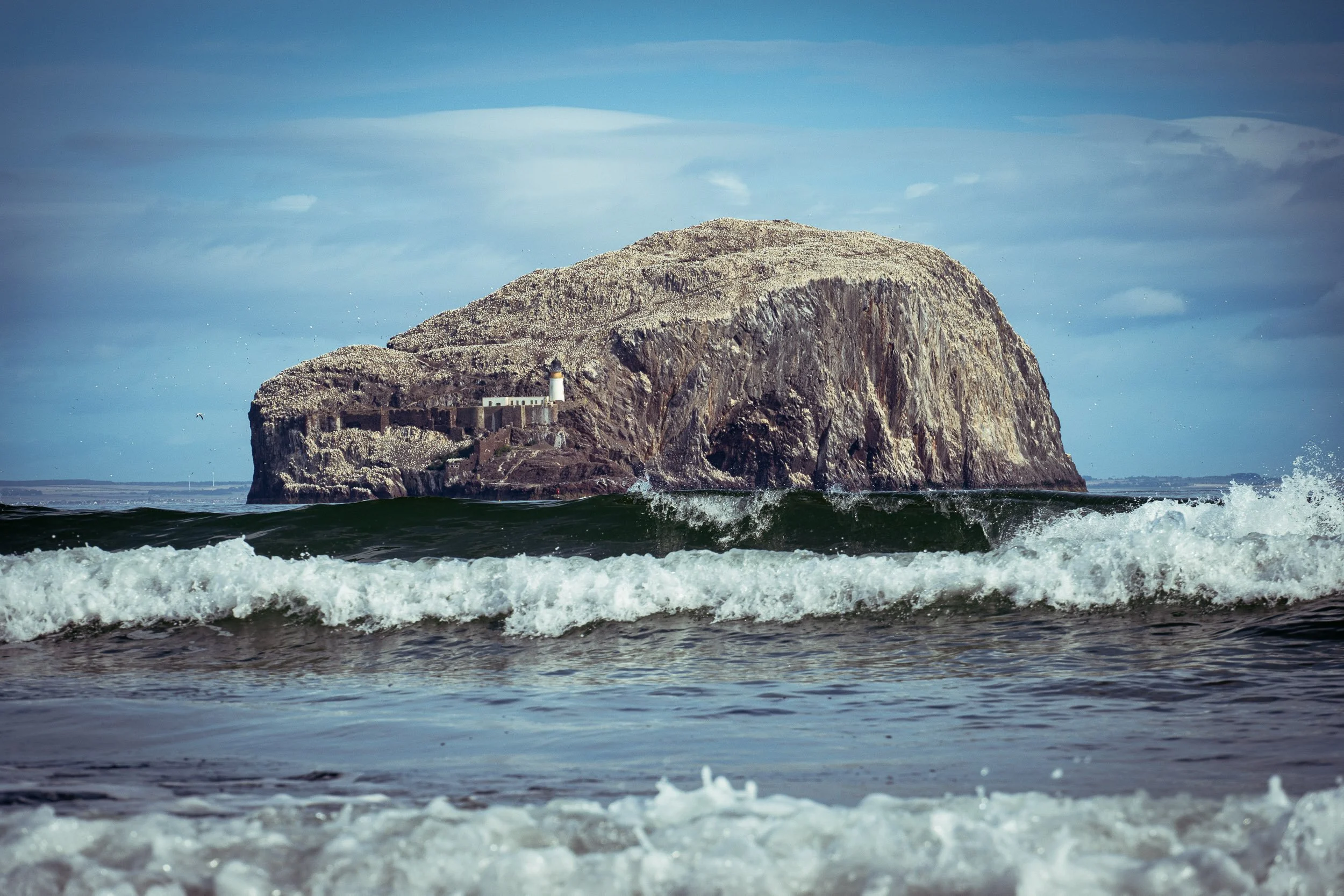 The Bass Rock off the coast of East Lothian, surrounded by ocean waves, birds and a partly cloudy sky.