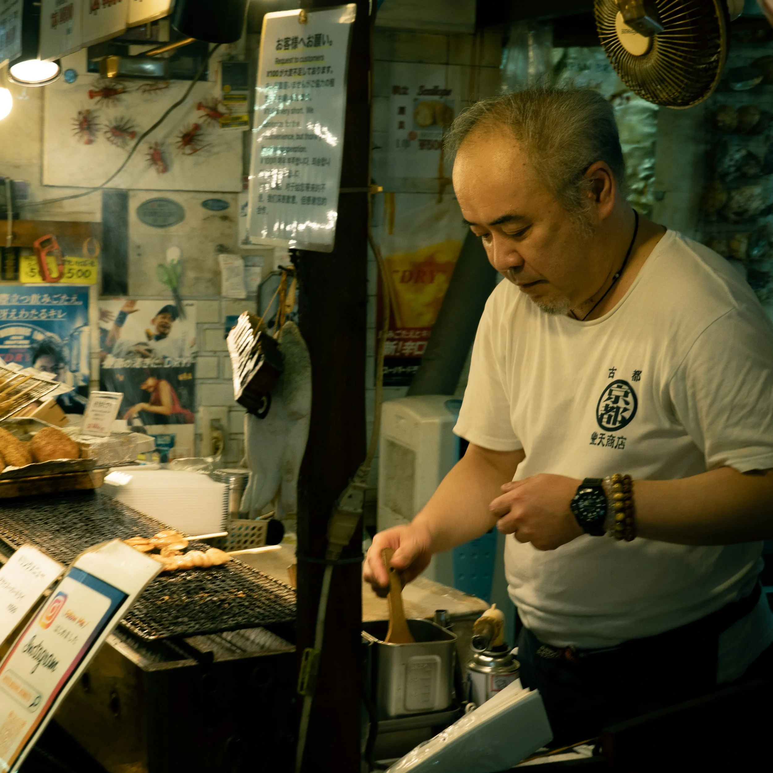 Man with gray hair and wearing a white T-shirt and black bracelets standing at a counter in a store, with various signs, coffee beans, and a fan visible in the background.