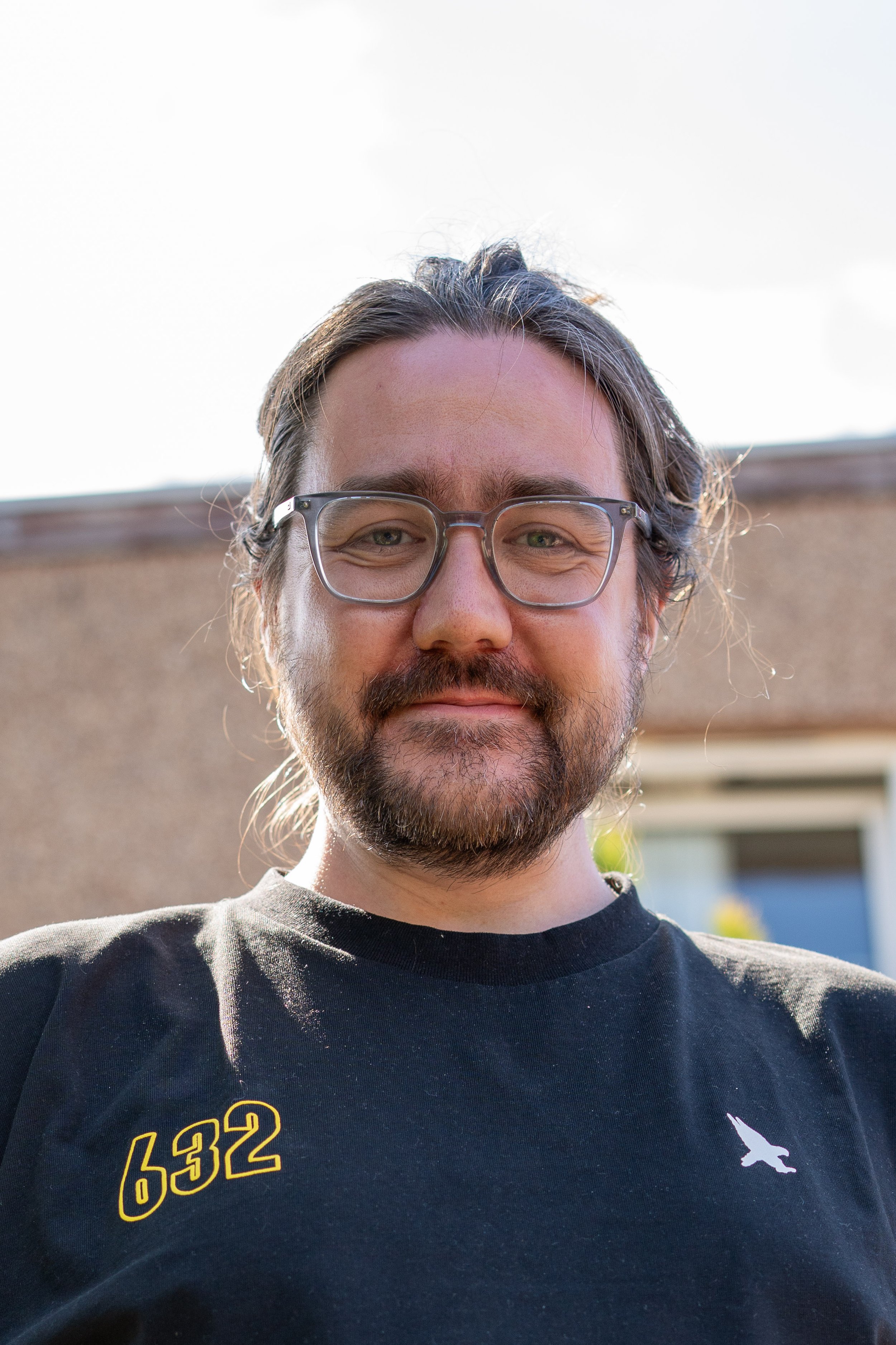 Grant Taylor, photographer. A man with glasses, long hair, and a beard, smiling outdoors with a building in the background. He is wearing a black shirt with yellow numbers and a white logo.