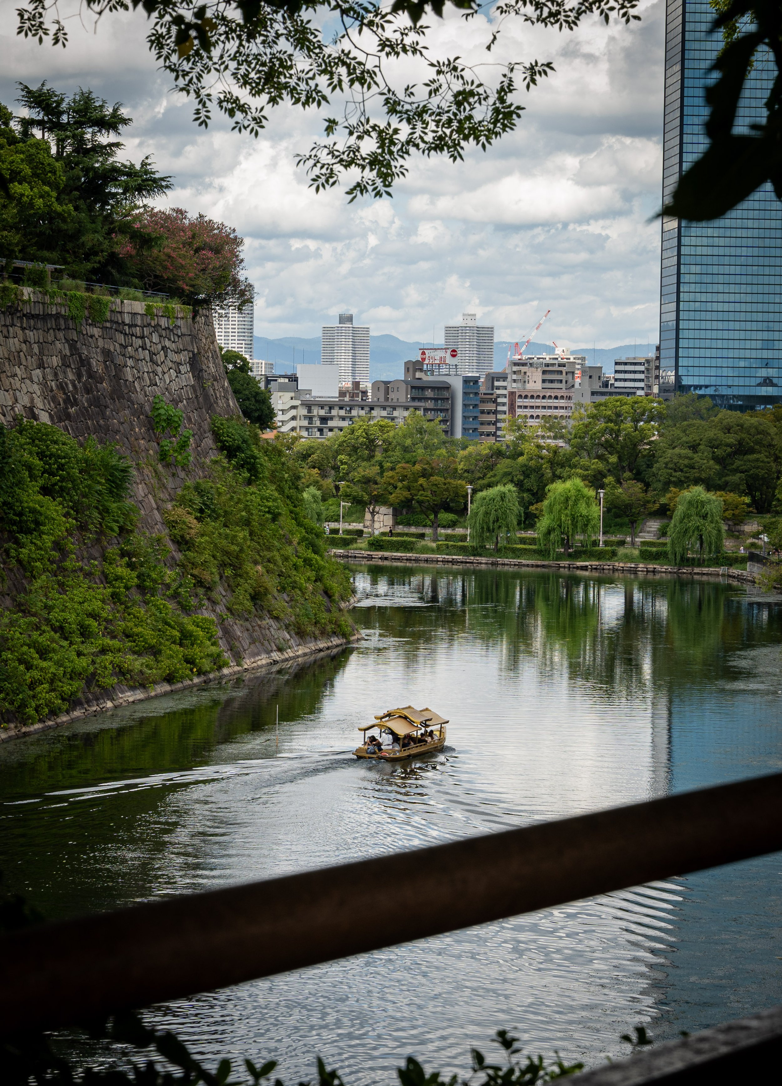 A small boat with people on it floating on the water surrounding Osaka Castle with city buildings and greenery in the background.