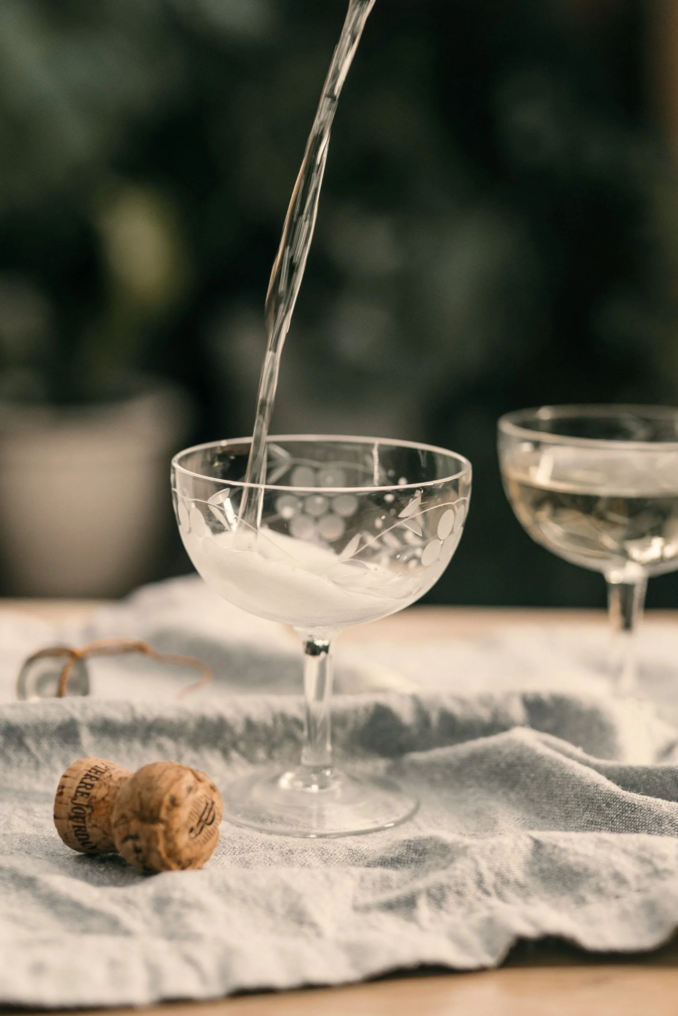 A close-up of a glass champagne coupe with polka dot pattern, being filled with a sparkling beverage. Two cork stoppers are visible on a white cloth underneath the glass.