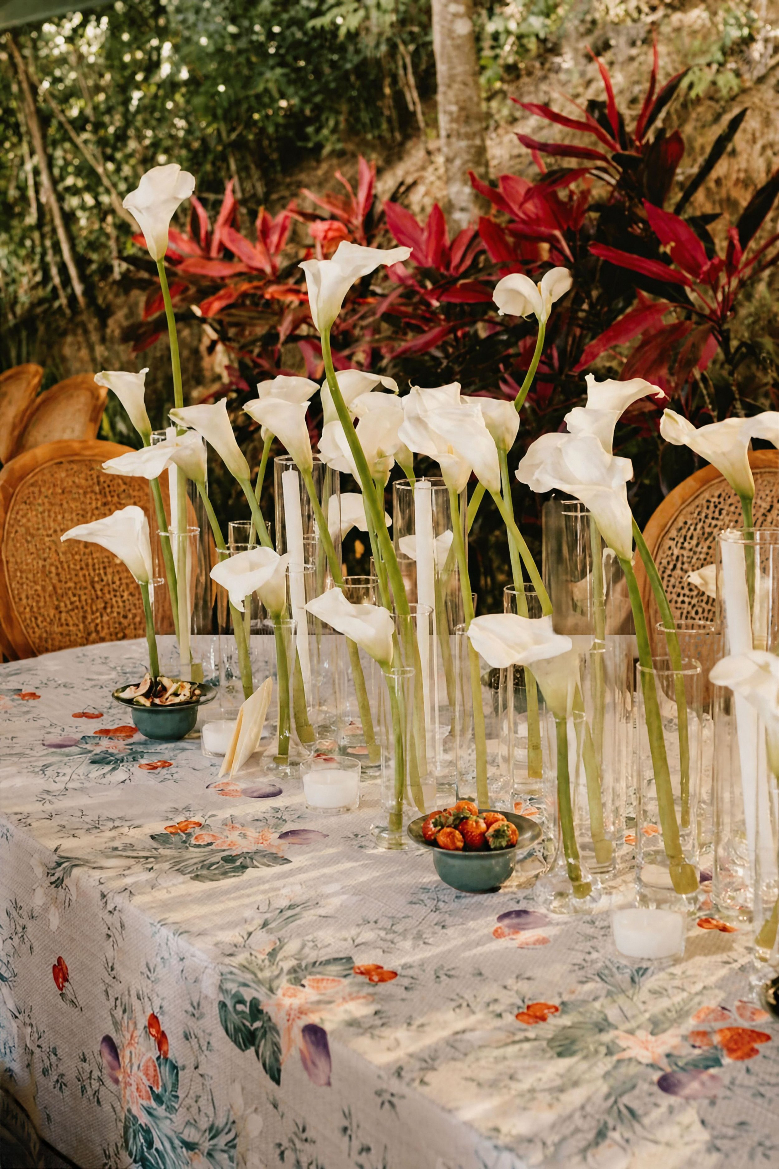 A table decorated with tall white calla lilies in glass vases and small teal bowls with strawberries, set in a floral tablecloth. In the background, there are chairs with woven backs and a garden with green and red foliage.