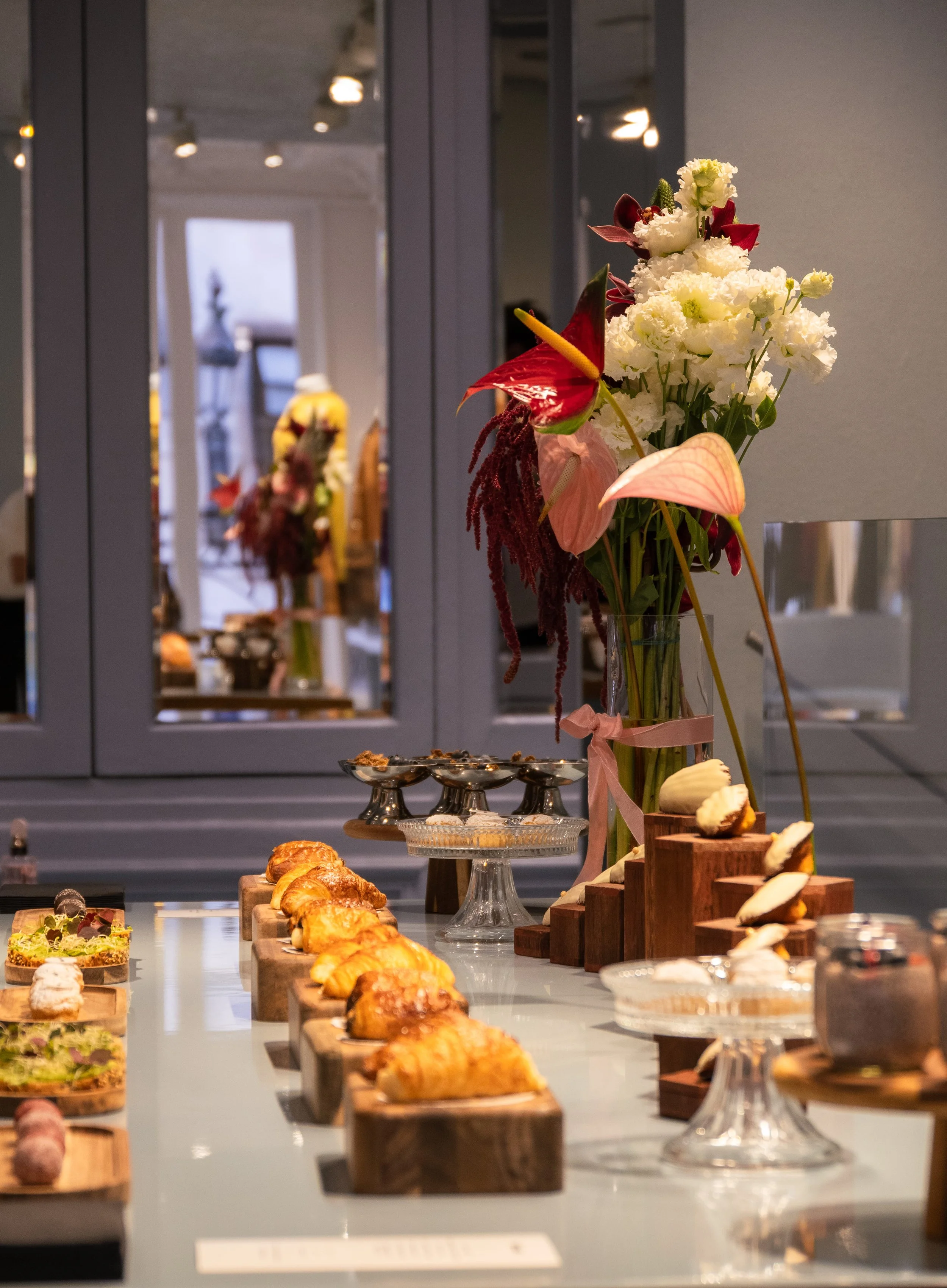 Une table avec une bouquet de fleurs colorées et diverses pâtisseries sur des supports en bois et en verre, dans une pièce lumineuse.