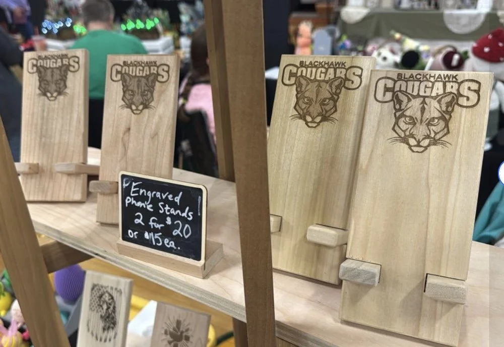 Wooden engraved phone stands featuring the Blackhawk Cougars logo with a cougar's face, displayed on a wooden shelf at a market stall.