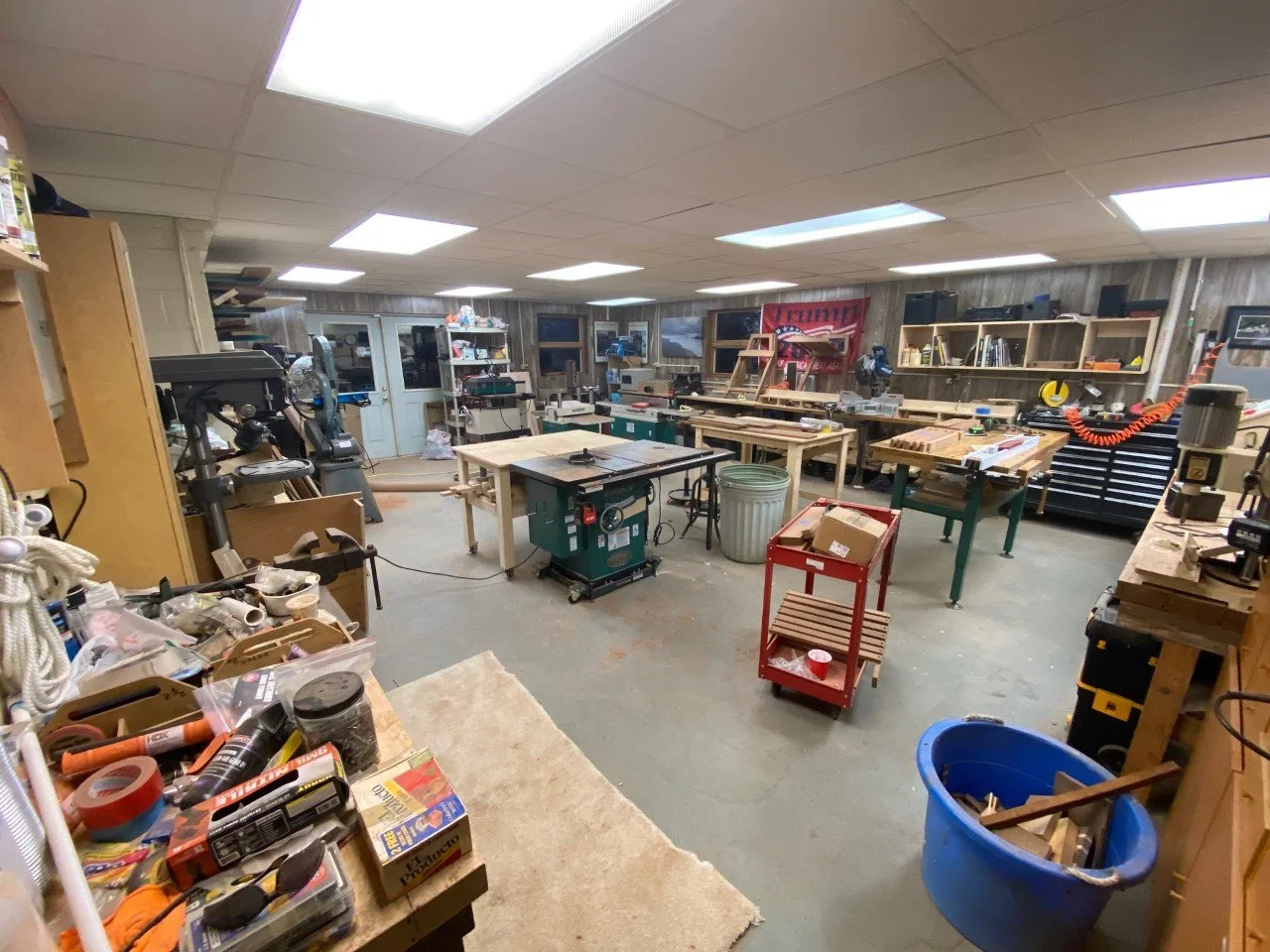 A cluttered woodworking workshop with tools, workbenches, shelves, and various construction materials.