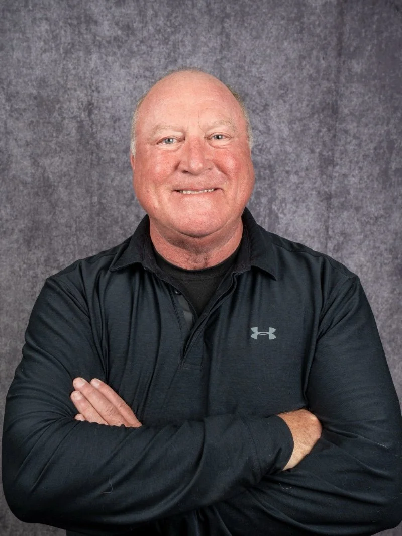A smiling middle-aged man with crossed arms, wearing a black Under Armour sports shirt, standing in front of a gray textured background.