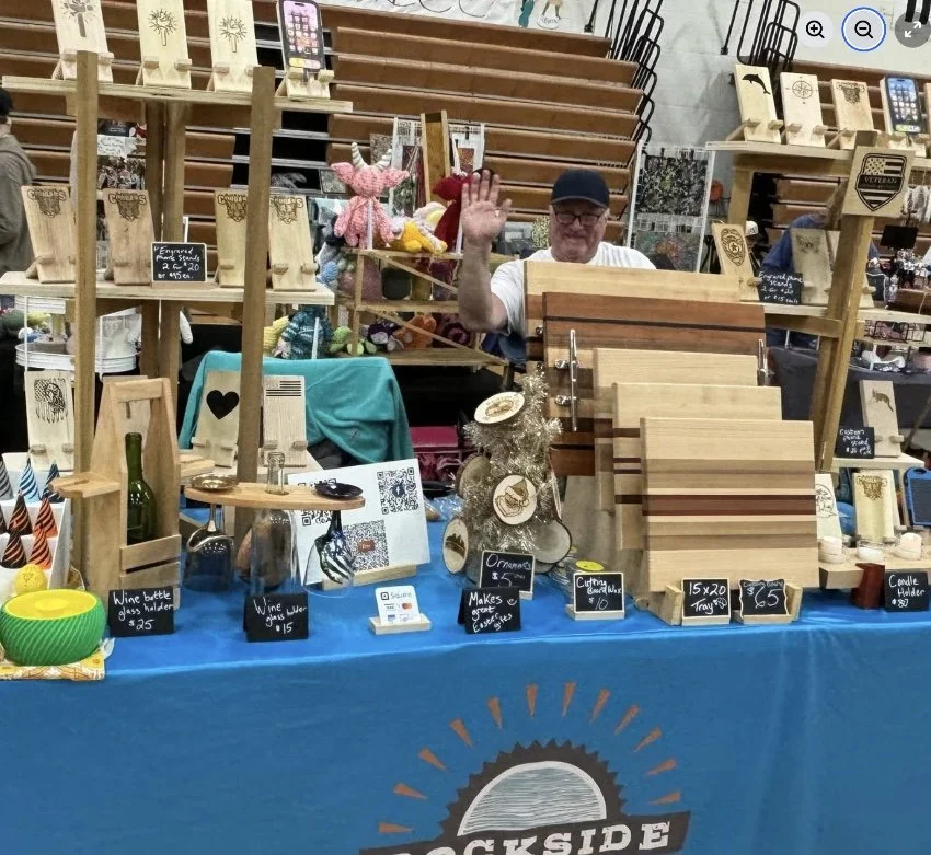 Man standing behind a table with handcrafted wooden cutting boards and other wood items at a craft fair.