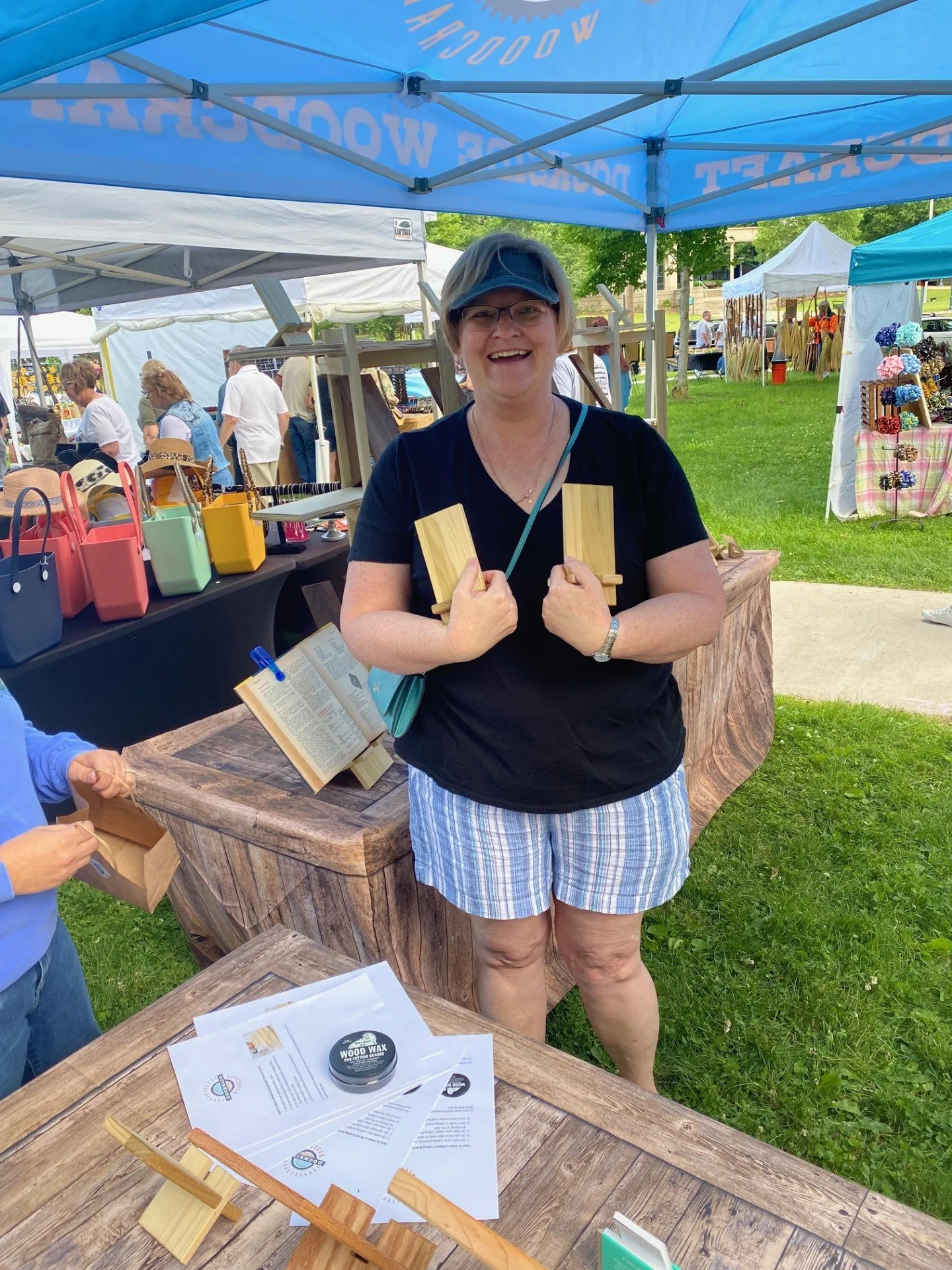 A smiling woman wearing glasses, a black shirt, and striped shorts holding two small wooden pieces at an outdoor market stall under a blue canopy. The stall has colorful bags, brochures, and wooden items displayed on the table. Other market booths and people browsing are visible in the background.