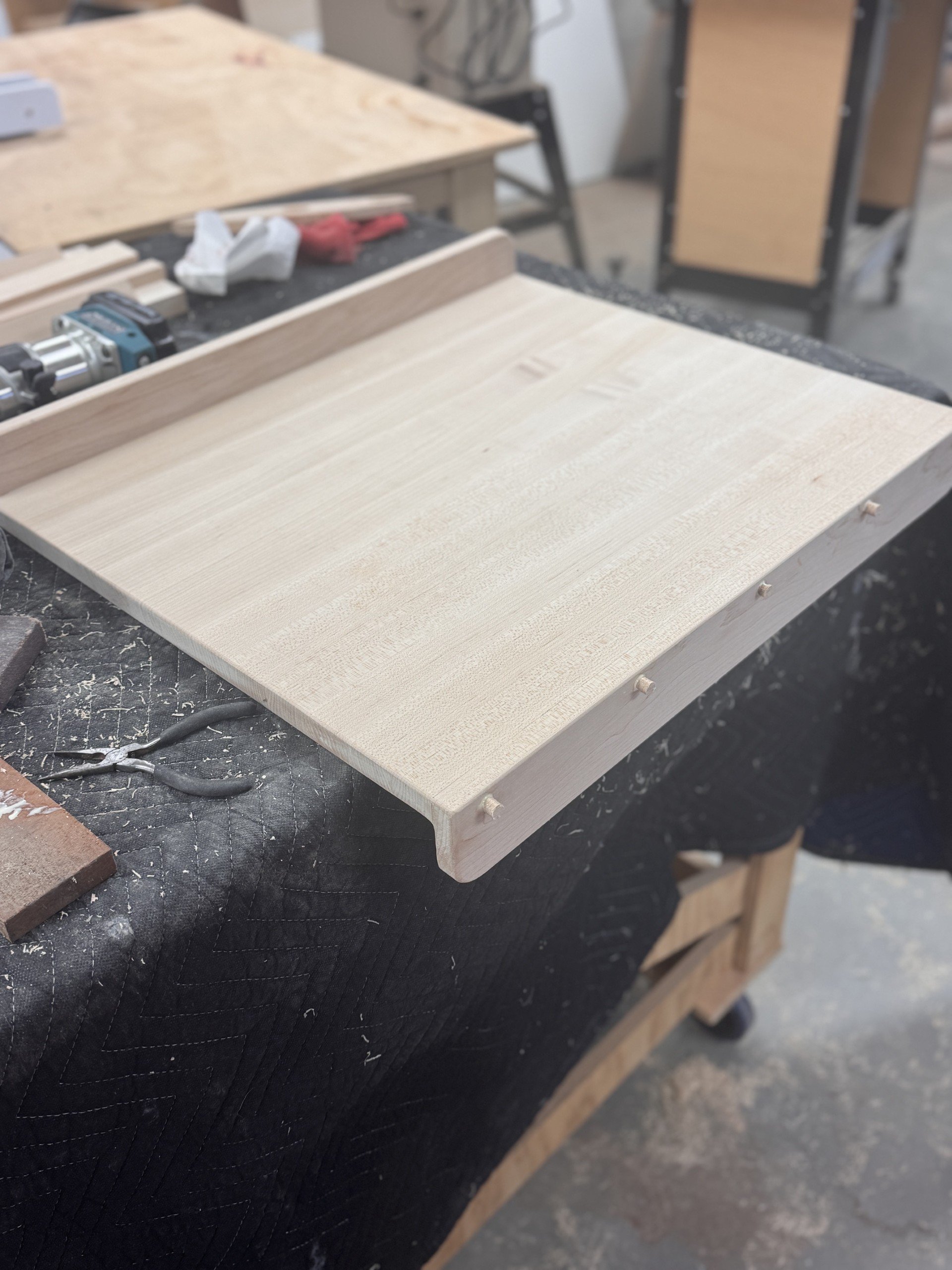 Close-up of a woodworking project on a workbench, with a light-colored wooden panel and a partially visible woodworking planer or router. The background shows workshop tools and furniture.