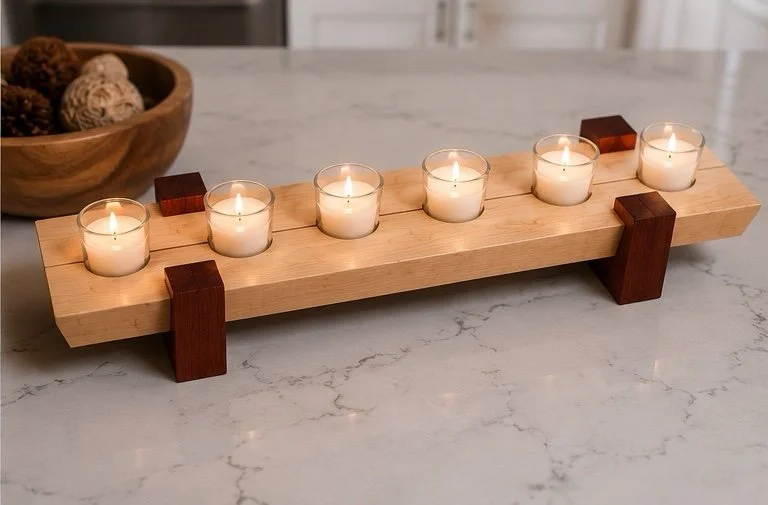 A wooden candle holder with six white tealight candles on a marble countertop, with a wooden bowl of decorative balls in the background.