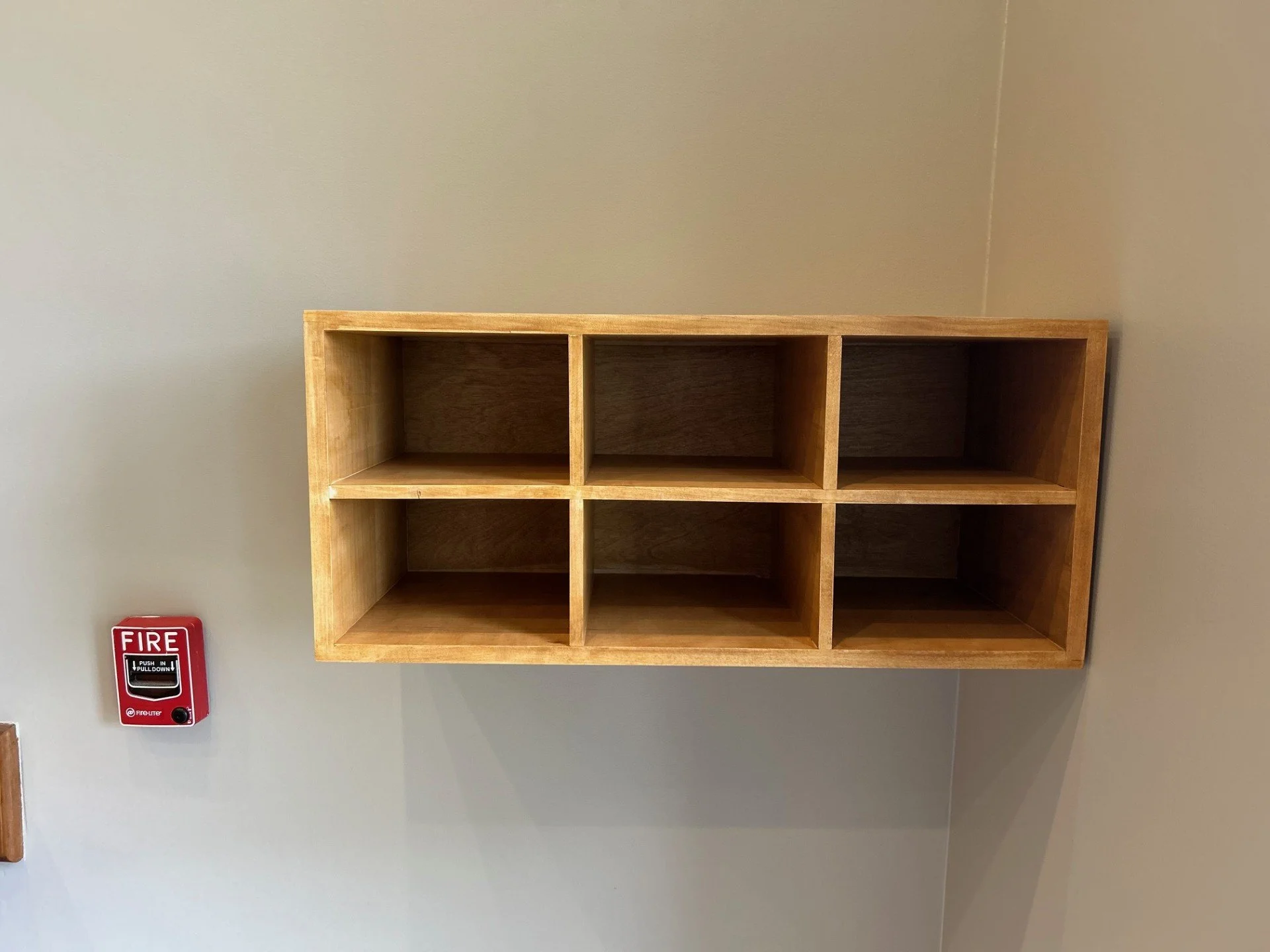 Wooden cubical shelf mounted on a light-colored wall with a red fire alarm pull station below it.