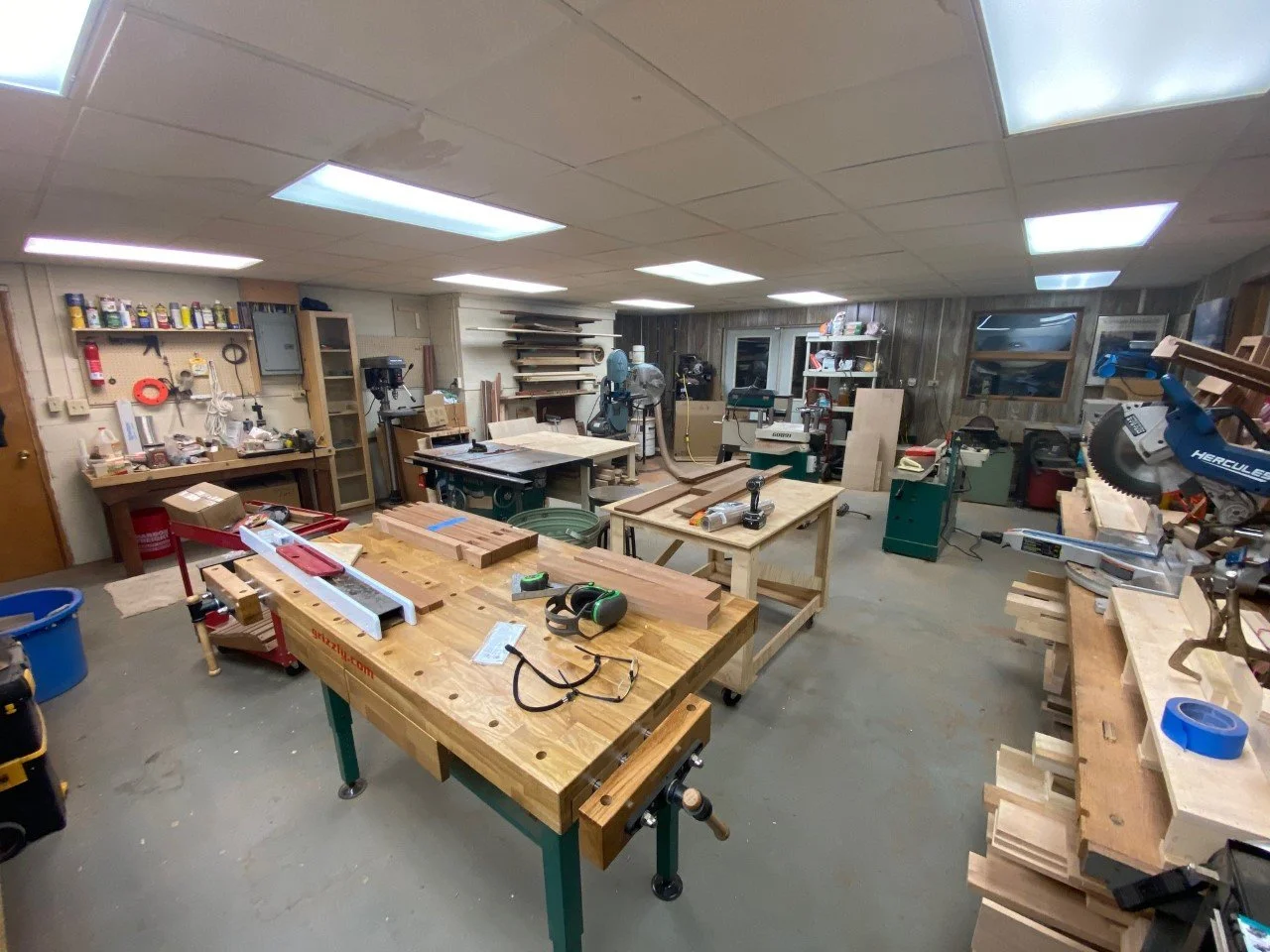 A woodworking workshop with tools, workbenches, and wood pieces, illuminated by fluorescent ceiling lights.