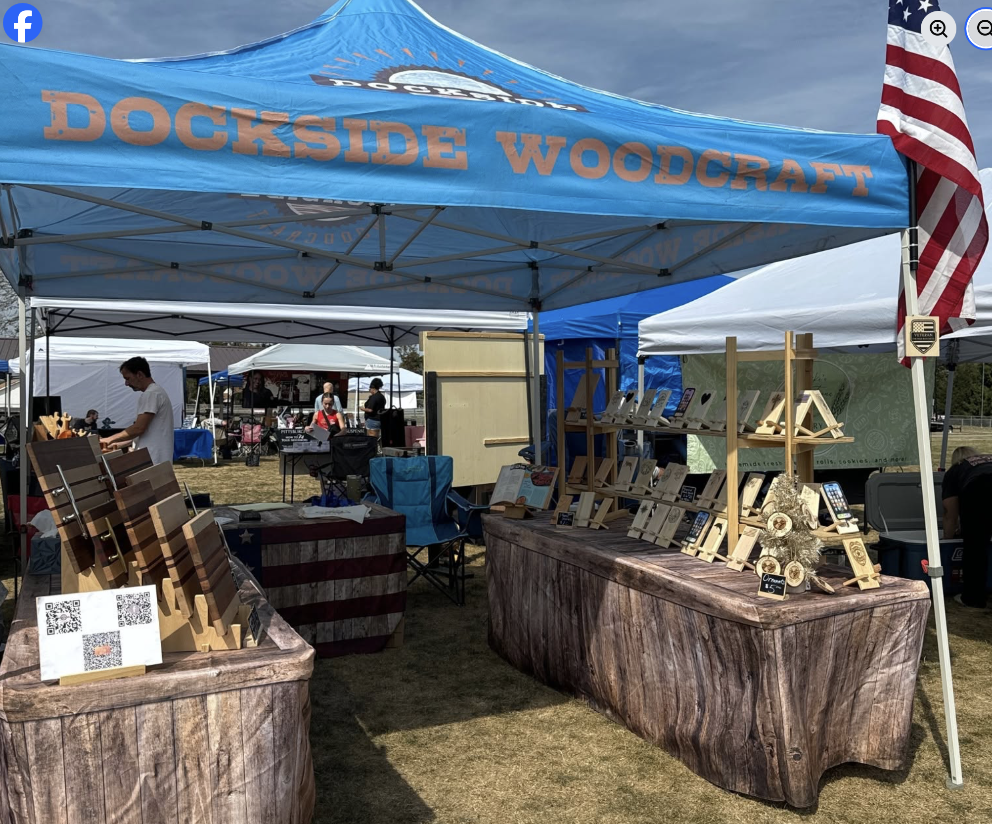 Outdoor market with booths, including one with a blue canopy reading 'Dockside Woodcraft,' and various wooden products on display, with American flag visible.