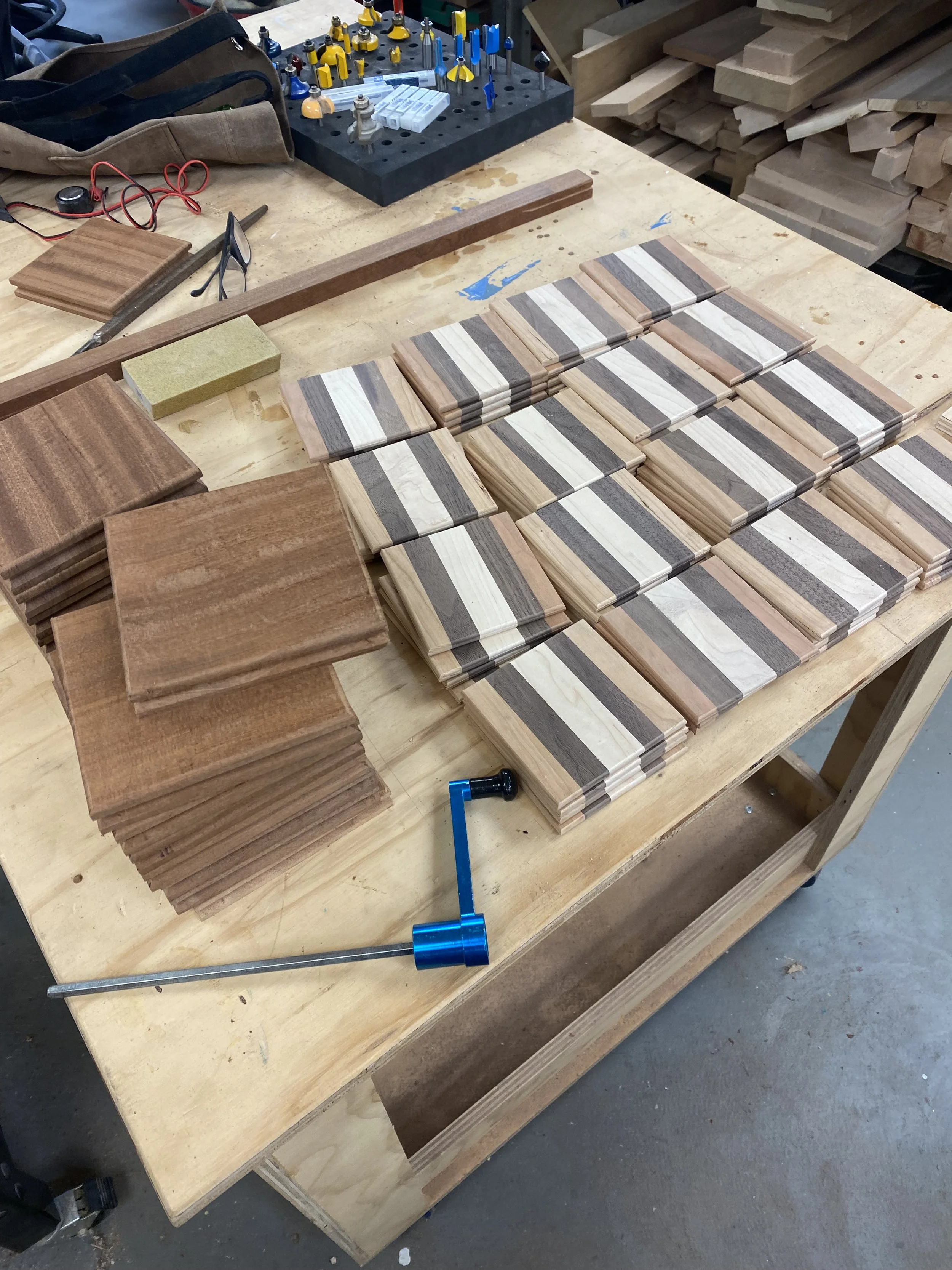 Various small wooden tiles with different wood grain patterns arranged on a workbench, with woodworking tools and supplies visible in the background.