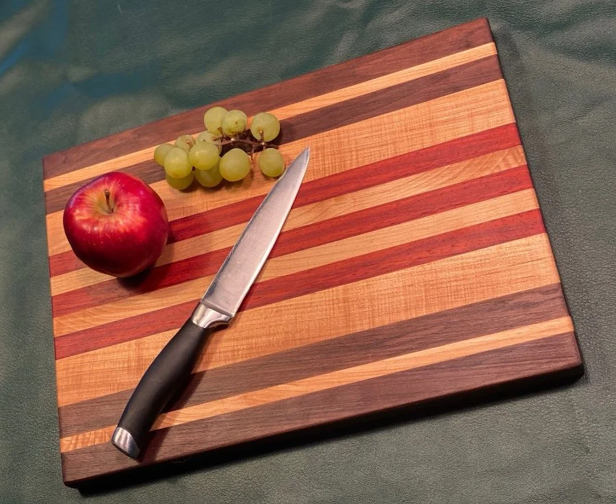 A wooden cutting board with red and light wood stripes, a knife, a red apple, and a small bunch of green grapes on it.