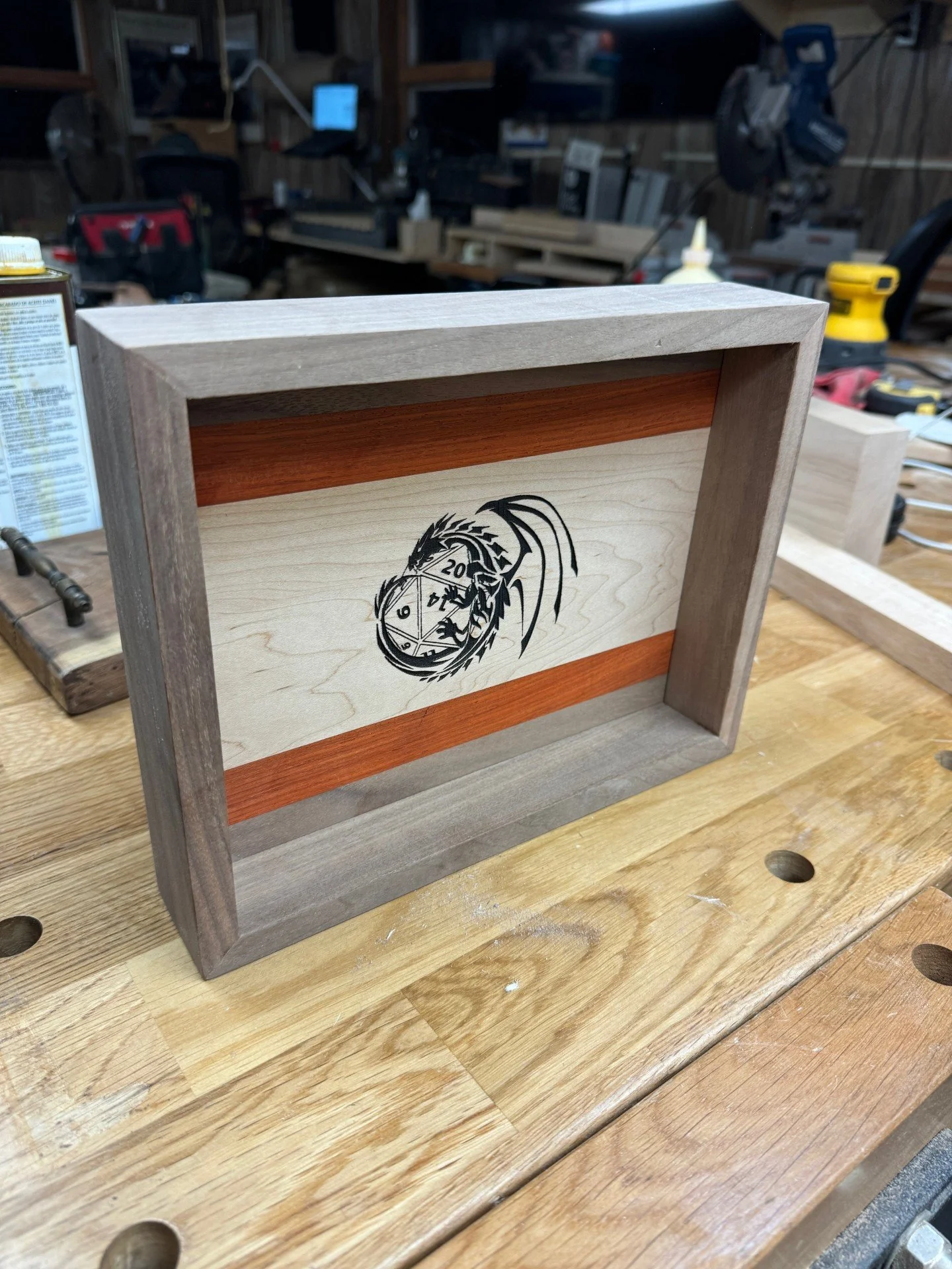 A wooden box with a dragon and D20 dice engraved on the front, sits on a workbench in a woodworking shop.