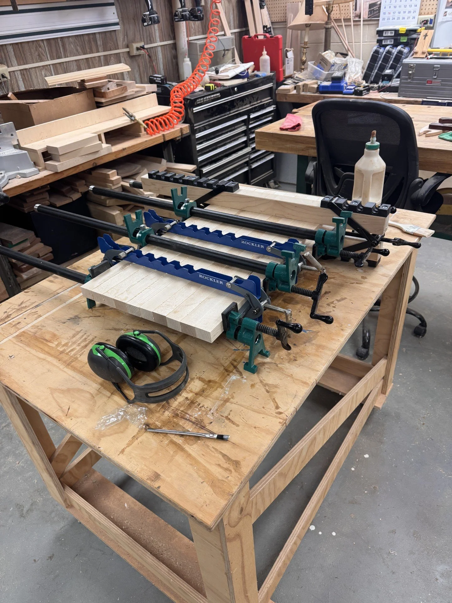 A woodworking workbench in a workshop with various tools and supplies. The workbench has clamps securing a piece of wood, a pair of green and black safety earmuffs, and a screwdriver. In the background, there are boxes, a chair, and shelves with more