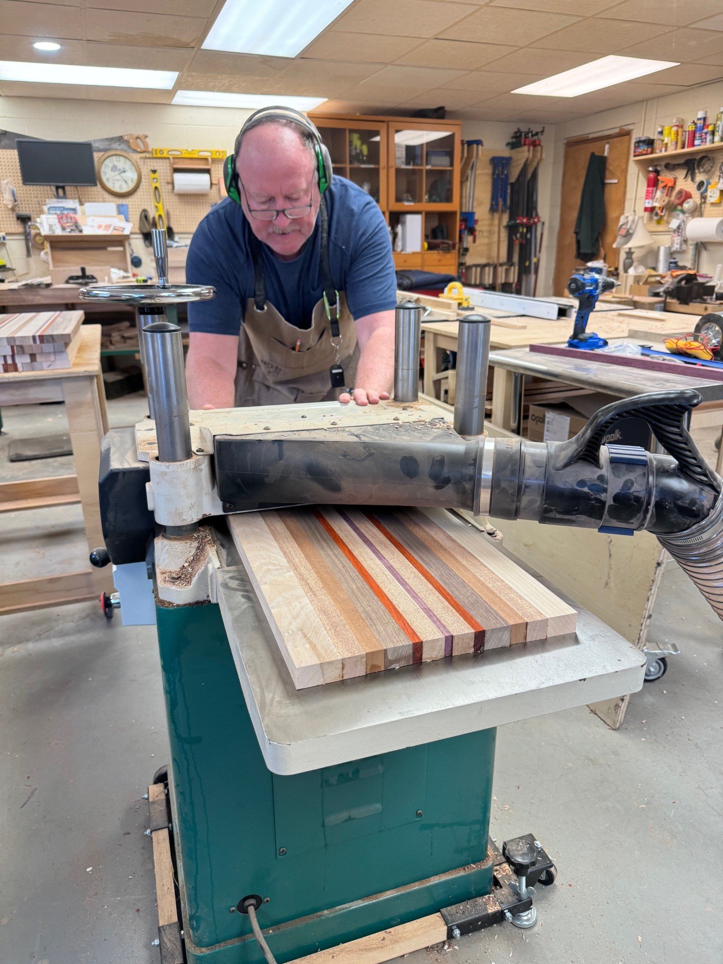 An older man wearing safety headphones and an apron, working on a woodworking project in a workshop with various tools and materials around him.