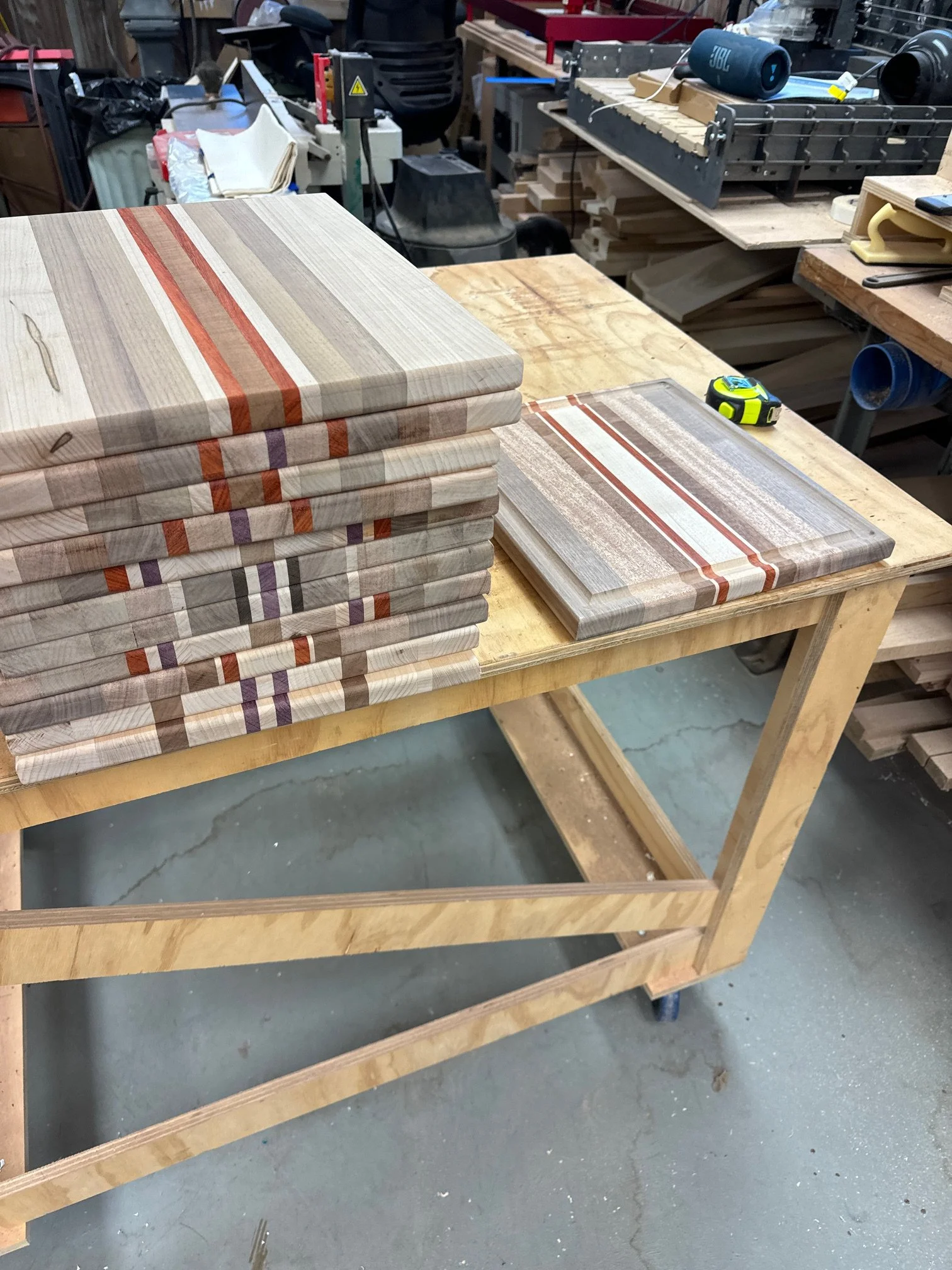 Stack of wooden cutting boards with colorful striped patterns on a workshop table, with woodworking tools and supplies visible in the background.