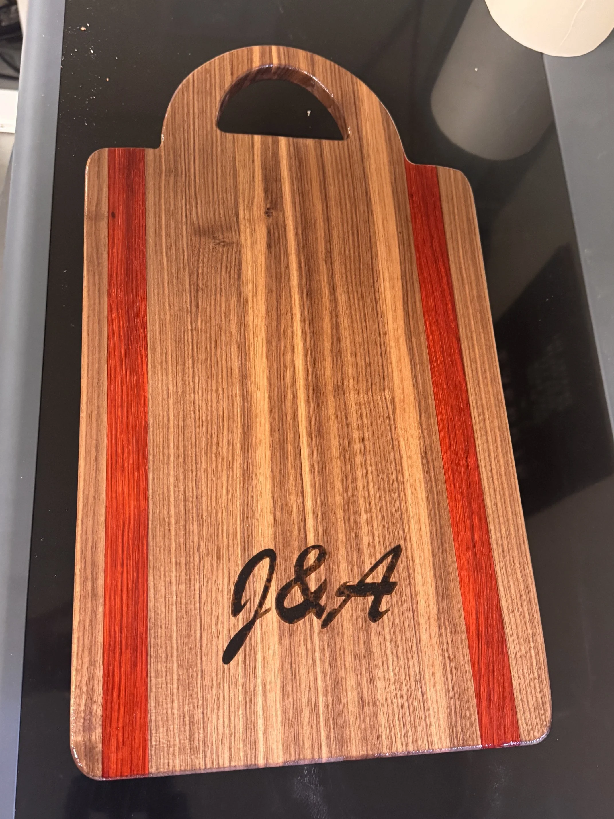 A wooden cutting board with a handle, placed on a black surface, with a white cup partially visible at the edge of the image.
