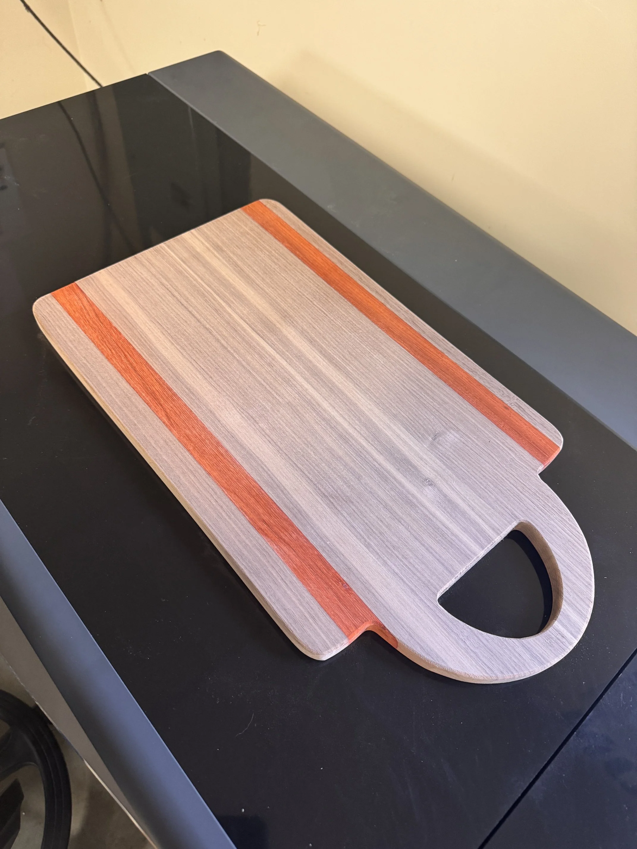 A rectangular wooden cutting board with a handle, placed on a black countertop. The board has a wood grain pattern and a pinkish-orange stripe along one edge.
