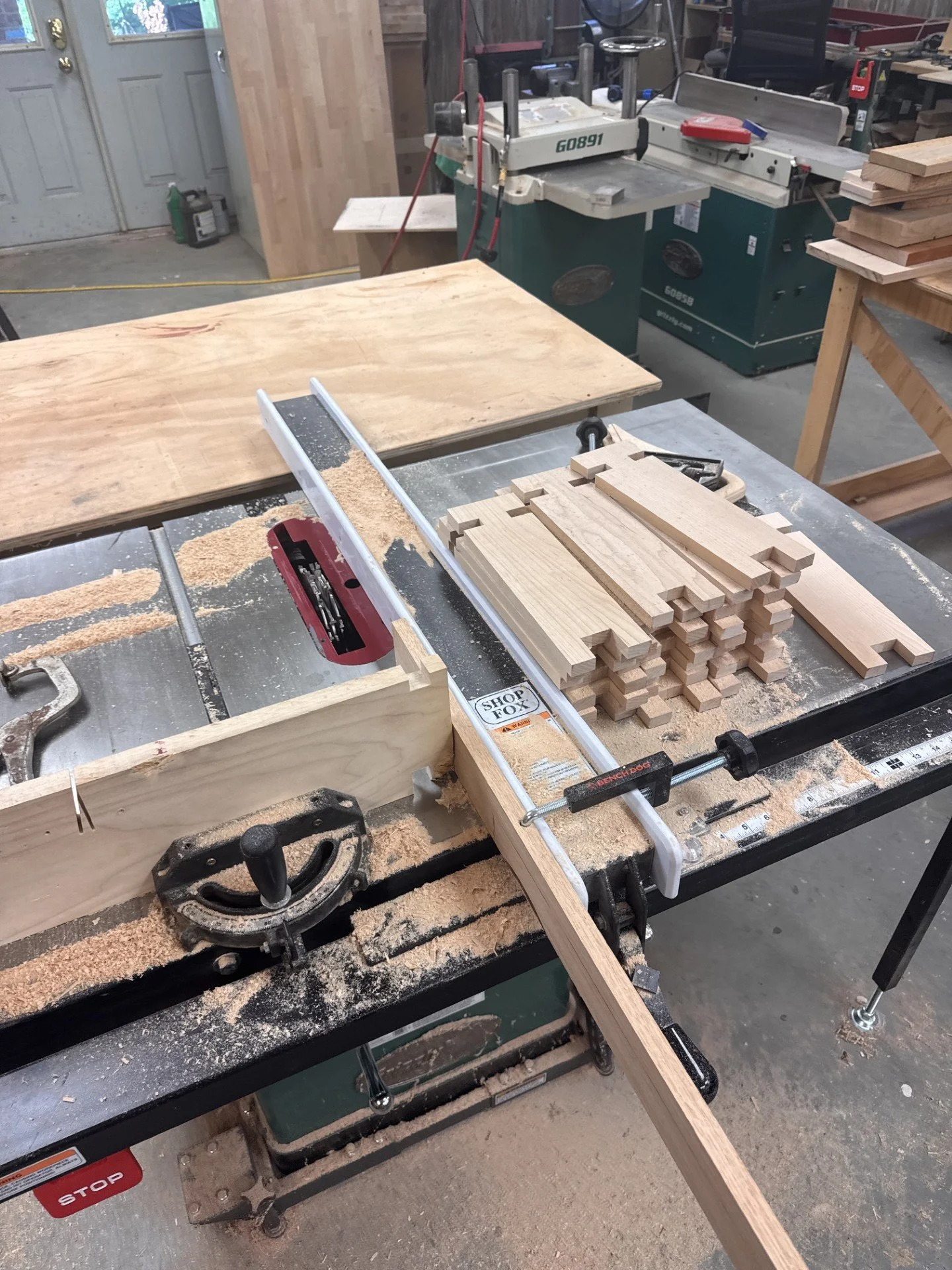 A woodworking workshop with a table saw, a miter saw, and a stack of cut wood pieces on a workbench.