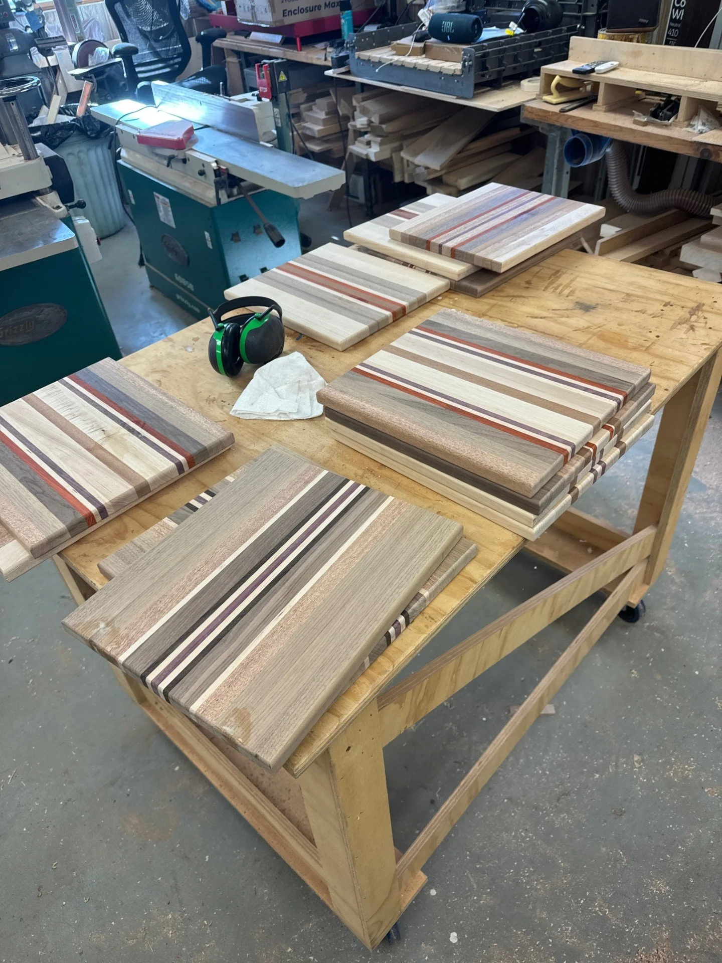 A woodworking workshop with a wooden workbench displaying several striped wooden cutting boards. In the background, there are various woodworking tools, machines, and stacks of wood.