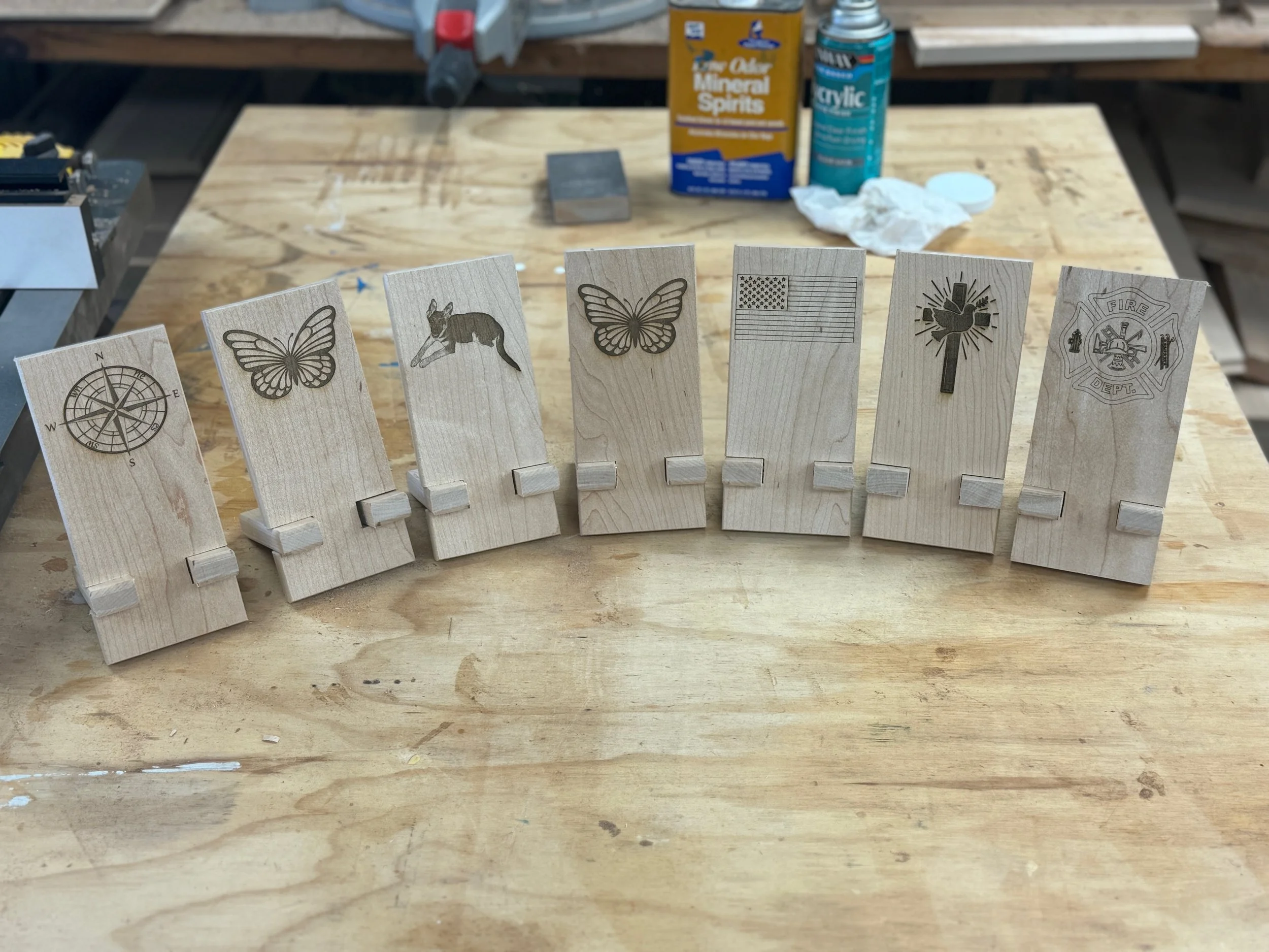 A row of wooden blocks decorated with engraved images, including a compass, butterfly, fox, American flag, dove with olive branch, and a firefighter badge, placed on a wooden table in a workshop setting with tools and supplies in the background.